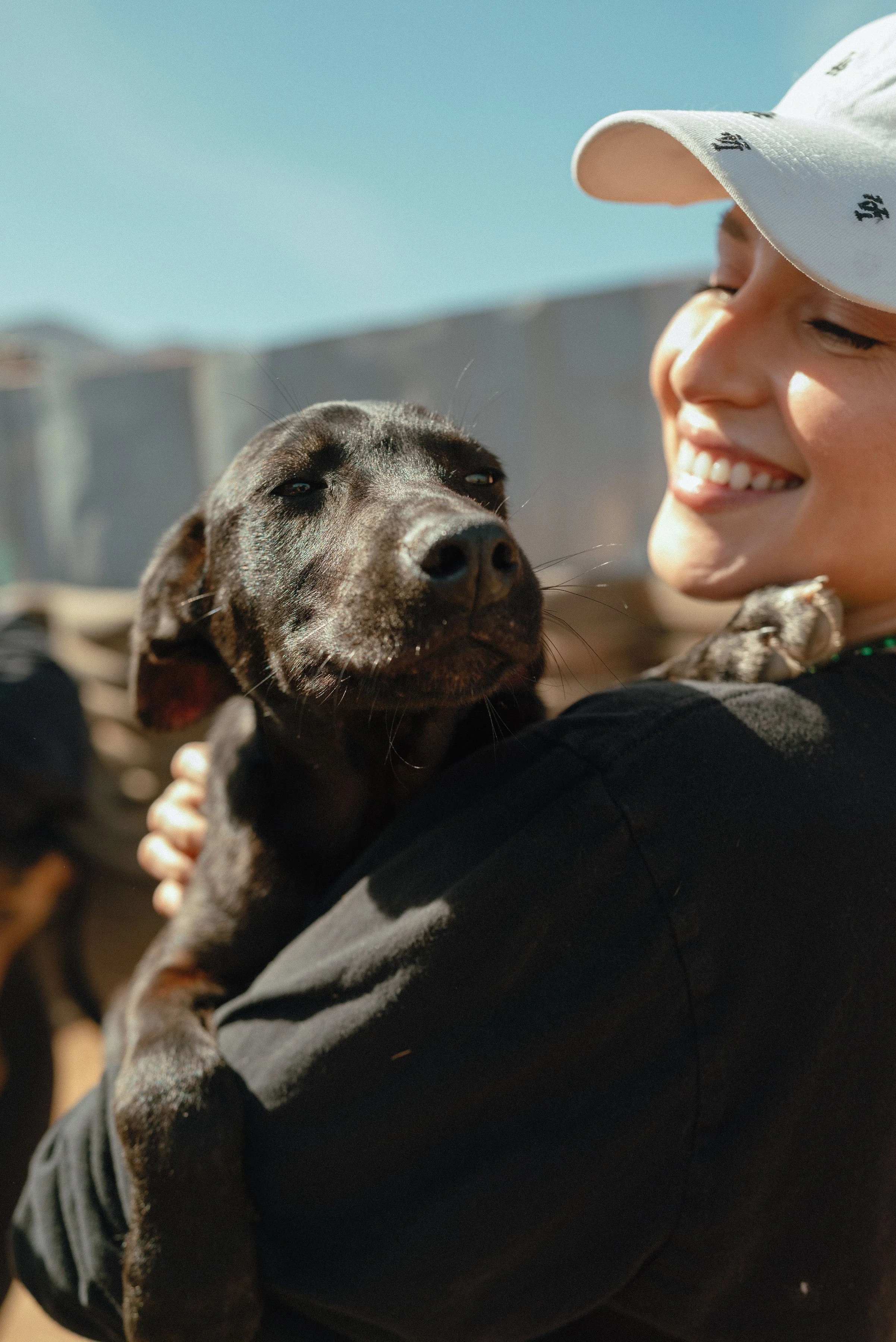 A smiling young woman wearing a white cap holding a puppy close to her face outdoors.