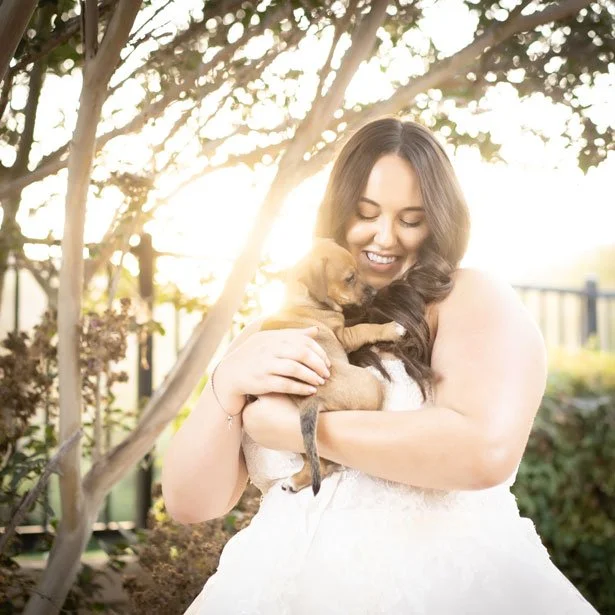 A woman in a white dress holding a small brown puppy in her arms, smiling and enjoying a sunny outdoor setting with trees and sunlight in the background.
