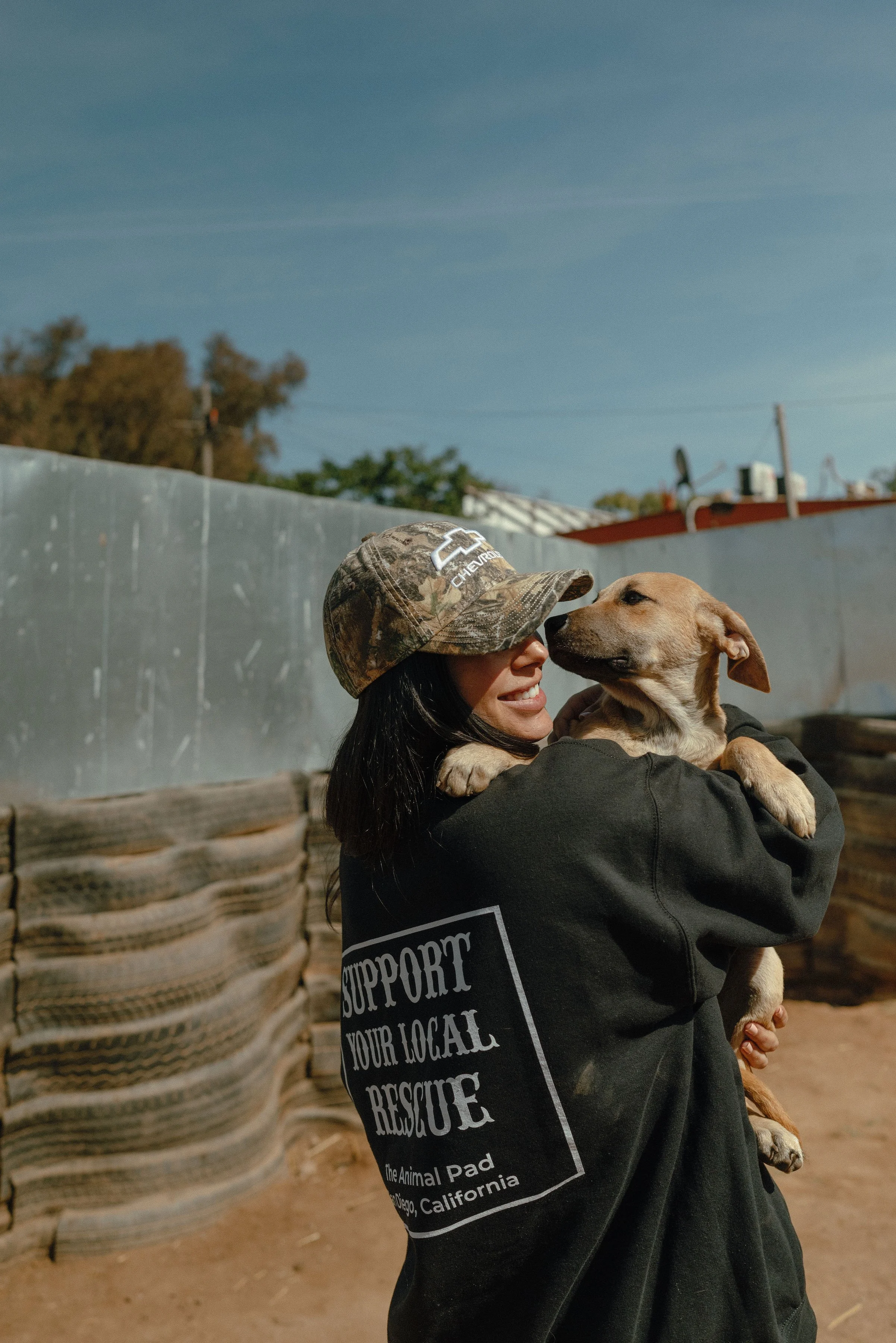 Person wearing a camouflage baseball cap holding a dog on their shoulder outdoors.