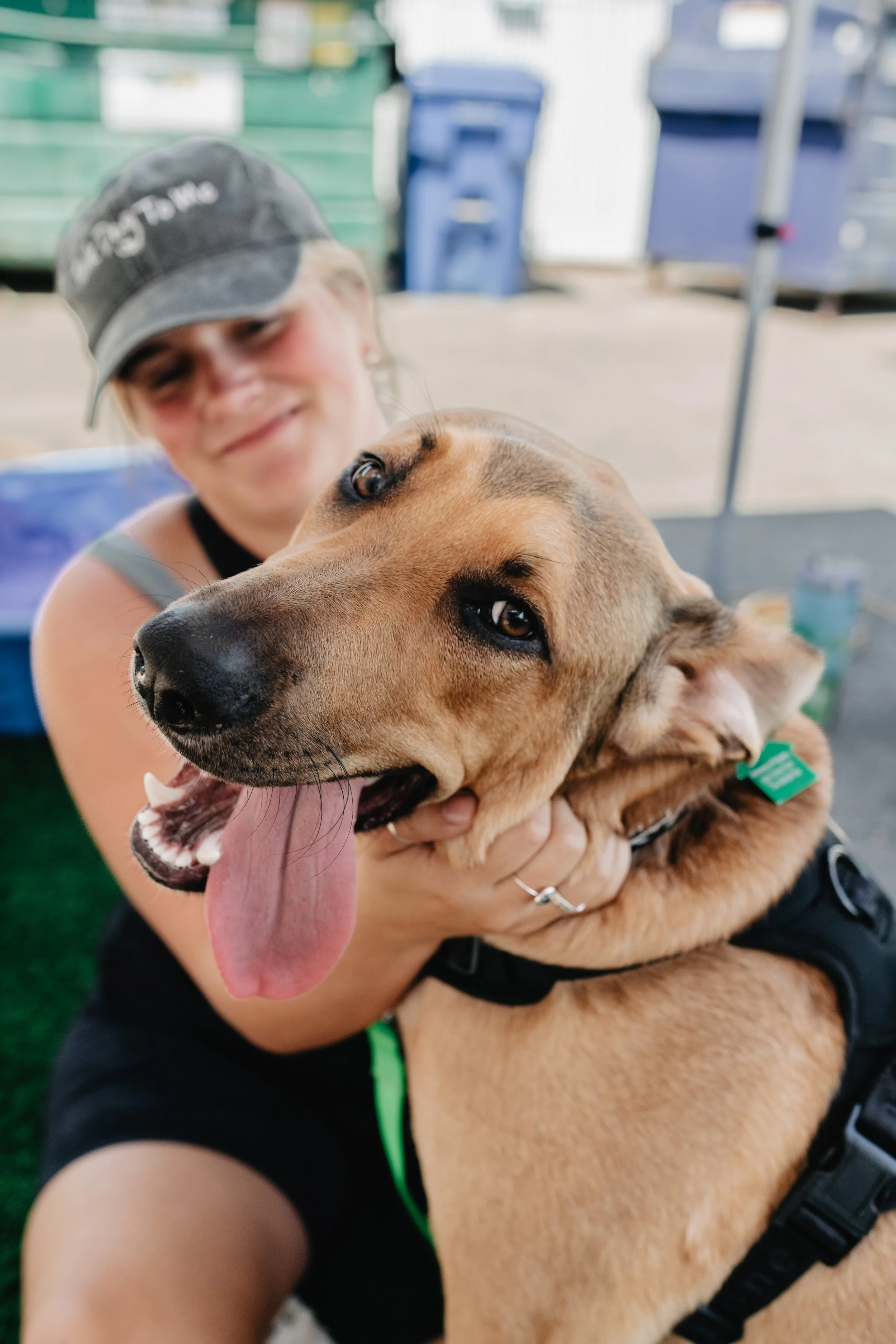 A woman wearing a gray cap with white text, smiling, holding a large tan dog with black markings and a black harness, inside a building with colorful trash bins in the background.