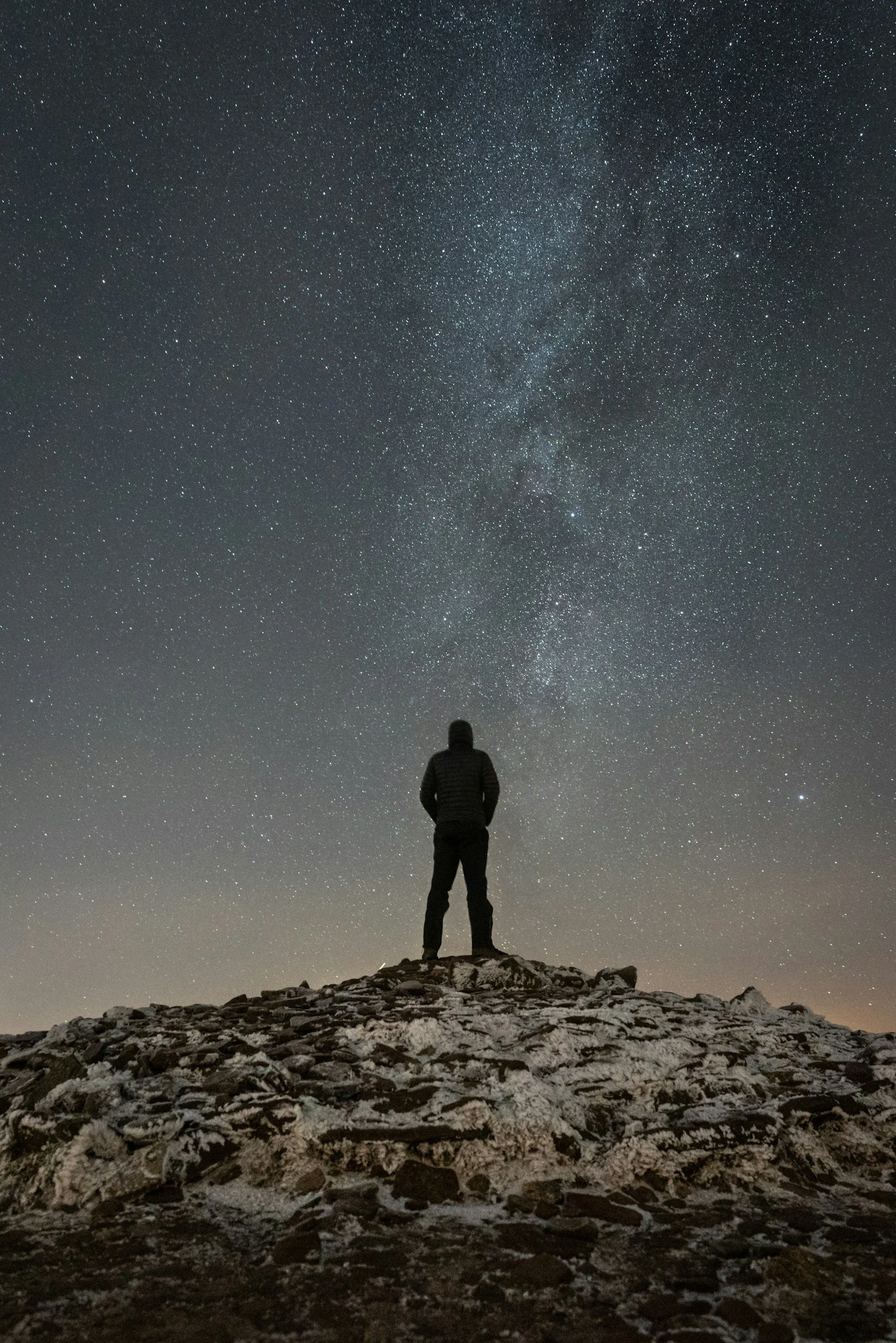 A person standing on a rocky hilltop at night, gazing at a star-filled sky with the Milky Way galaxy visible.