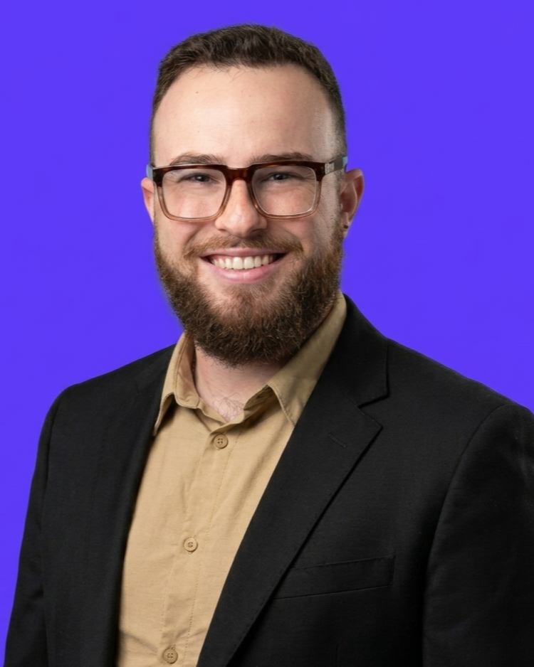 A smiling man with glasses, a beard, wearing a beige shirt and black blazer, against a purple background.