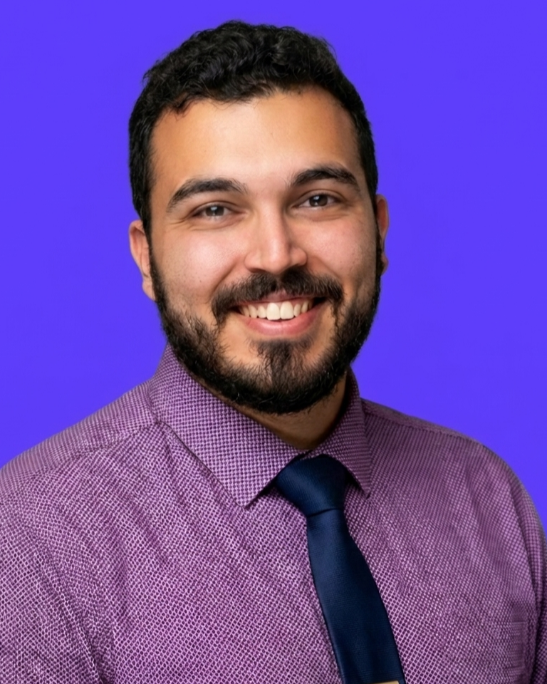 A professional headshot of a man with short dark hair and a beard, smiling, wearing a purple checked shirt and a navy blue tie, against a blue background.