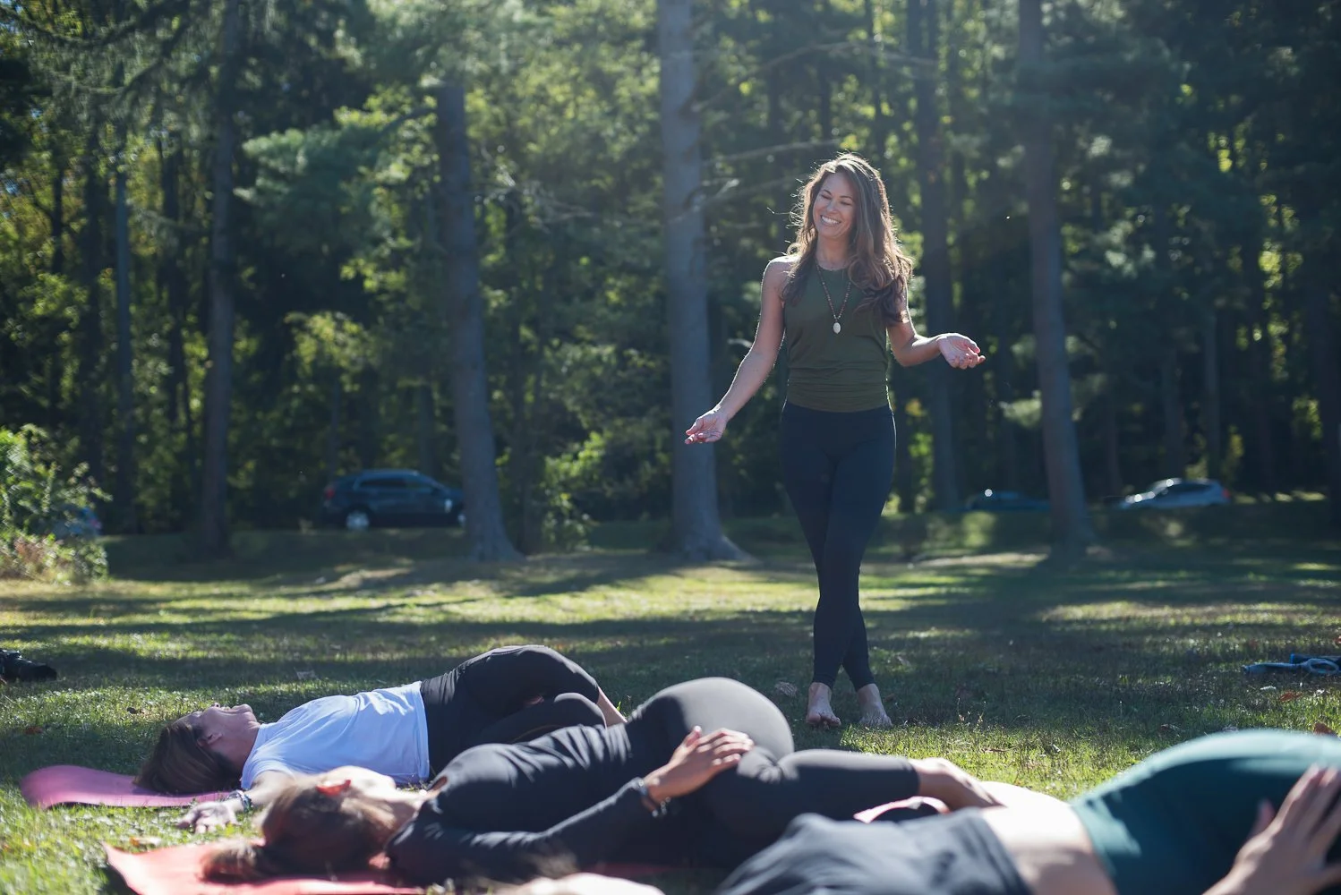 A yoga teacher standing outdoors on grass, smiling, participating in instructing yoga and or meditation class with friends at a park.