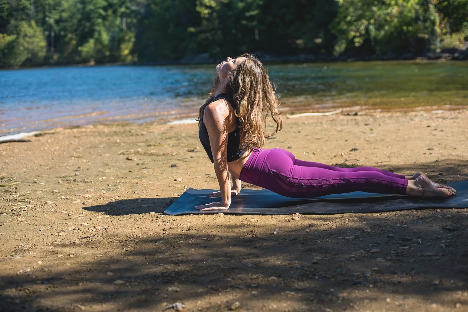 A woman practicing yoga outdoors on a mat by a river, performing a cobra pose with her face turned upwards, wearing purple leggings and a black top, surrounded by trees and natural scenery.