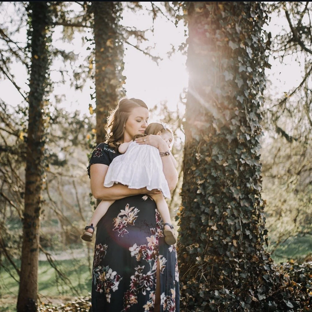 A woman with brown hair wearing a black floral dress holding a young girl in a white dress against a background of trees with sunlight filtering through.
