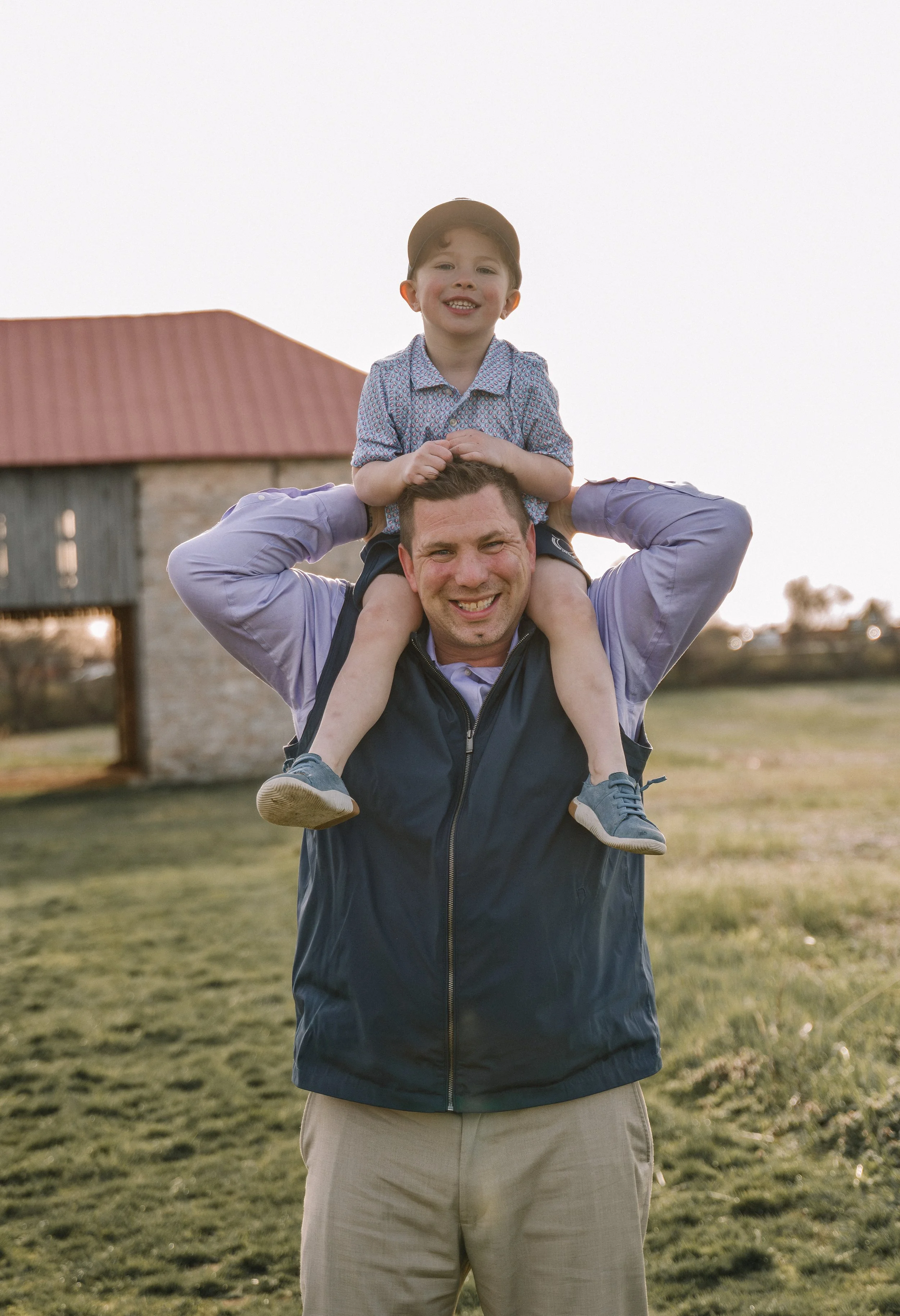 A man outdoors carrying a young boy on his shoulders, both smiling, with a barn and open field in the background during sunset.