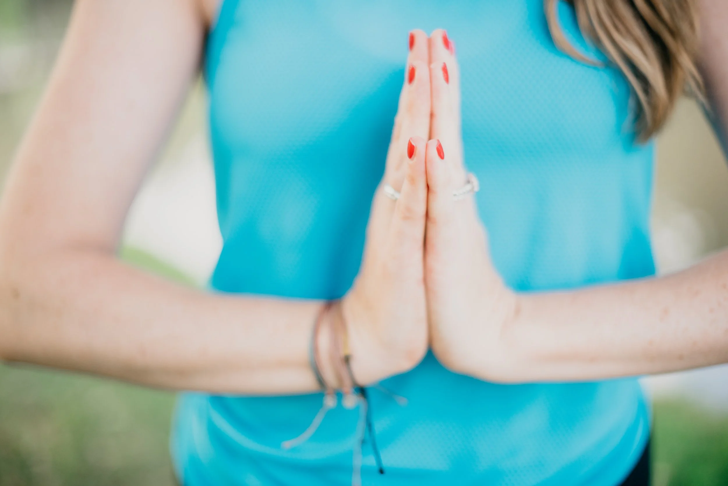 Close-up of a woman practicing yoga, standing outdoors, hands pressed together in prayer pose, wearing a blue athletic top, red nail polish, and rings, with a natural background.
