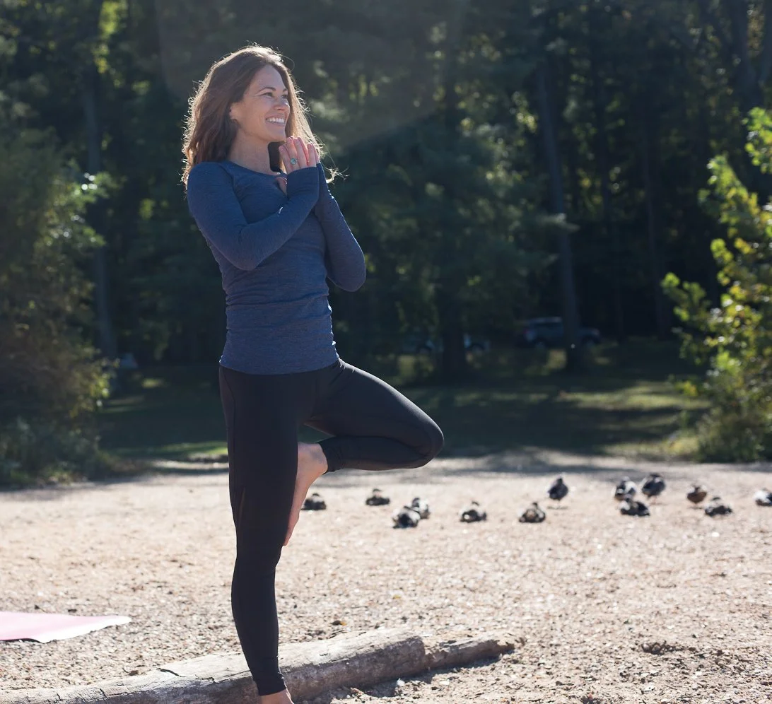 Woman practicing yoga outdoors in tree-lined park, standing on one leg in tree pose, smiling, wearing a long-sleeve athletic top and leggings.