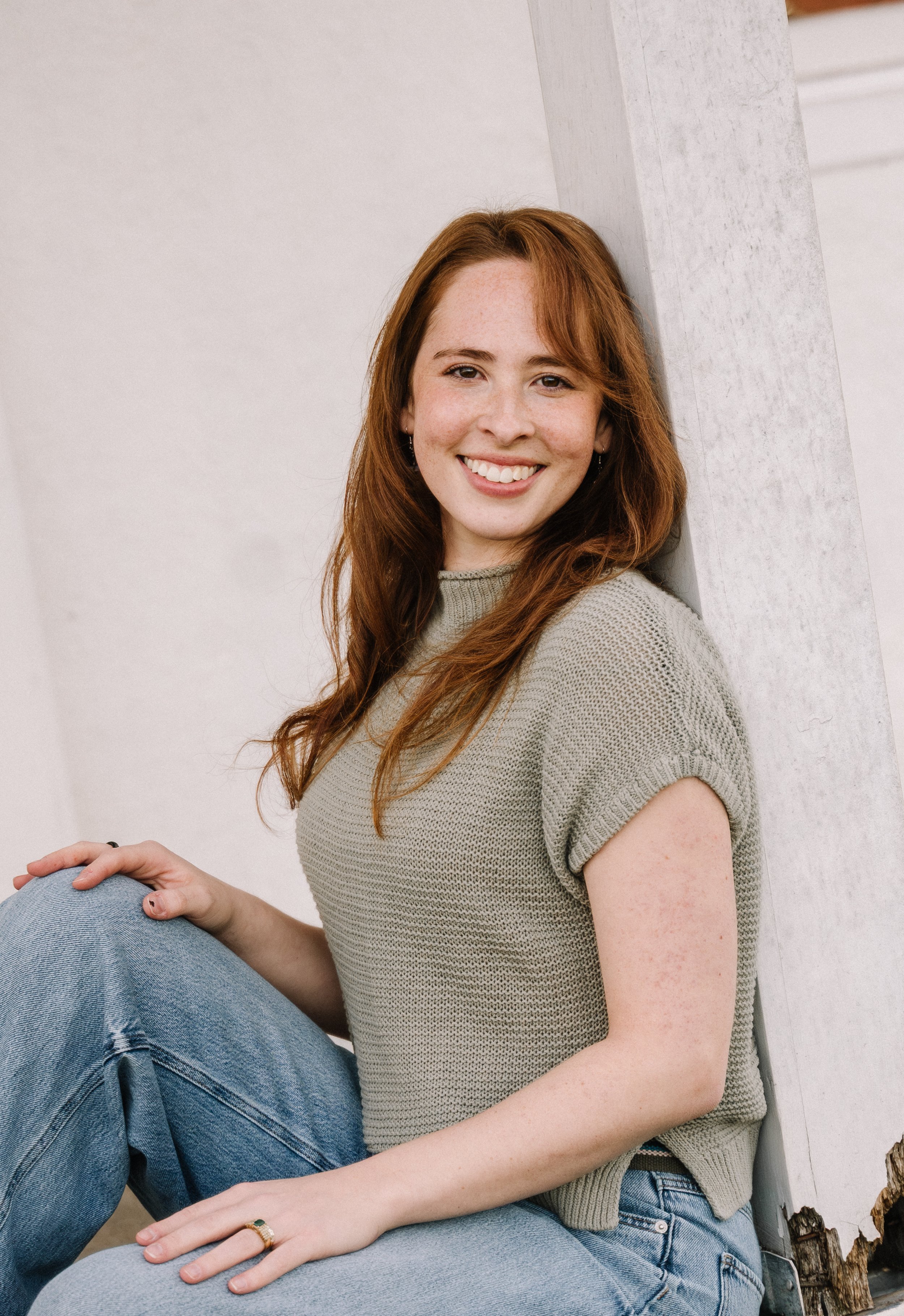 A young woman with long red hair and freckles, smiling, wearing a beige knit top and blue jeans, sitting on the ground leaning against a white wall, outdoors.