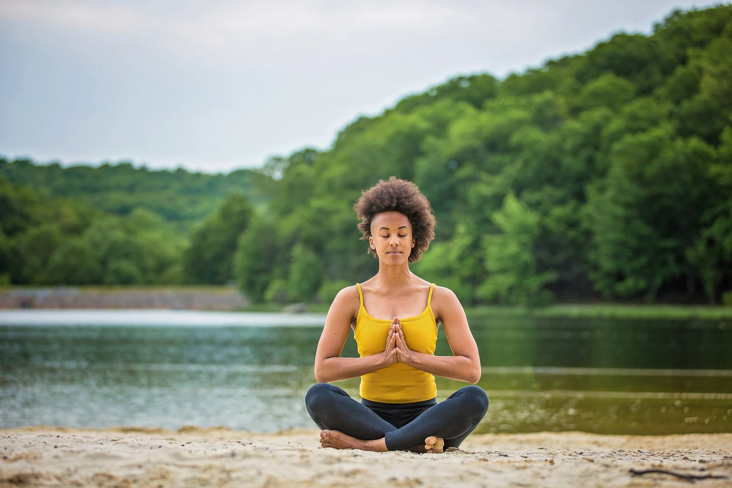 Woman meditating in a seated yoga pose on a sandy riverbank with green trees and water in the background.