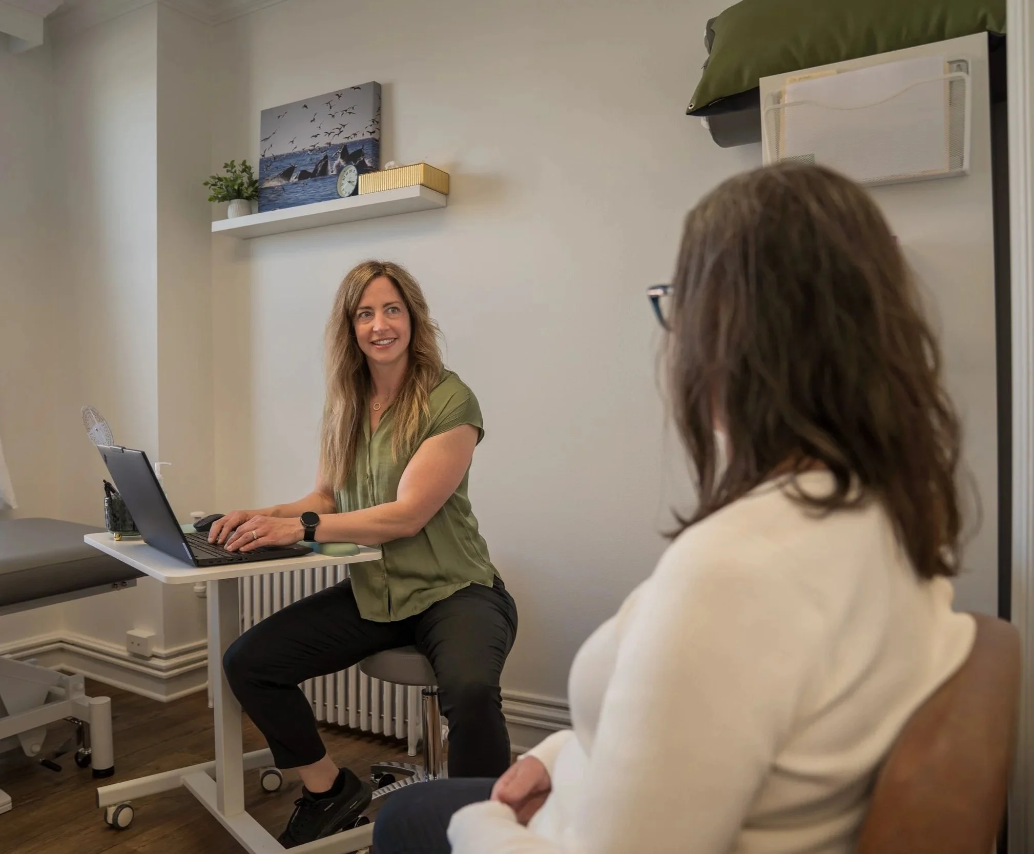 Two women having a conversation in an office or meeting room with a white wall background, a decorated shelf with a sunset seascape painting, a clock, and a plant, one woman is sitting at a desk with a laptop, and the other is sitting on a chair facing her.