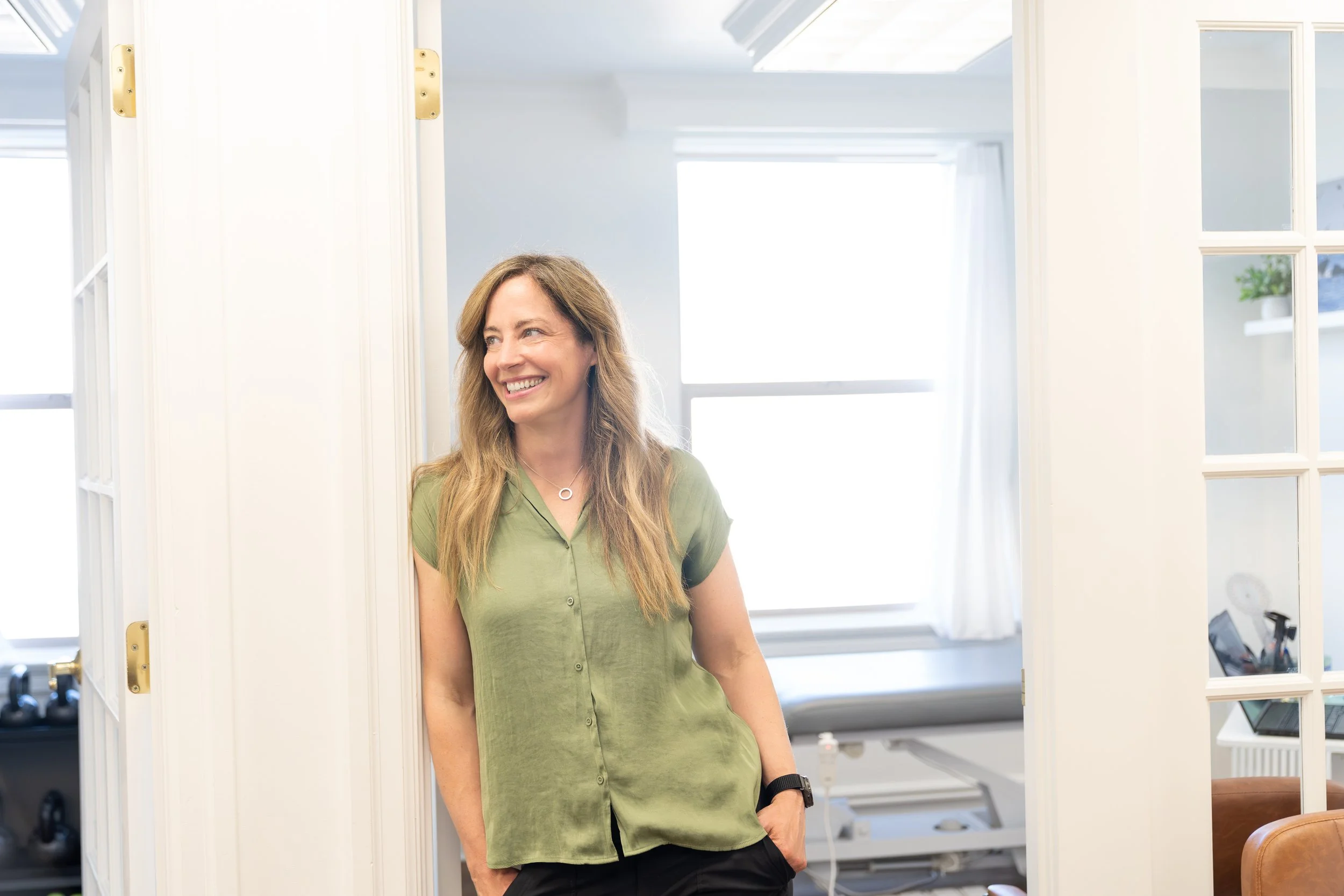 A woman with long light brown hair, wearing a green shirt, standing in a bright indoor setting, smiling, with her hand in her pocket.