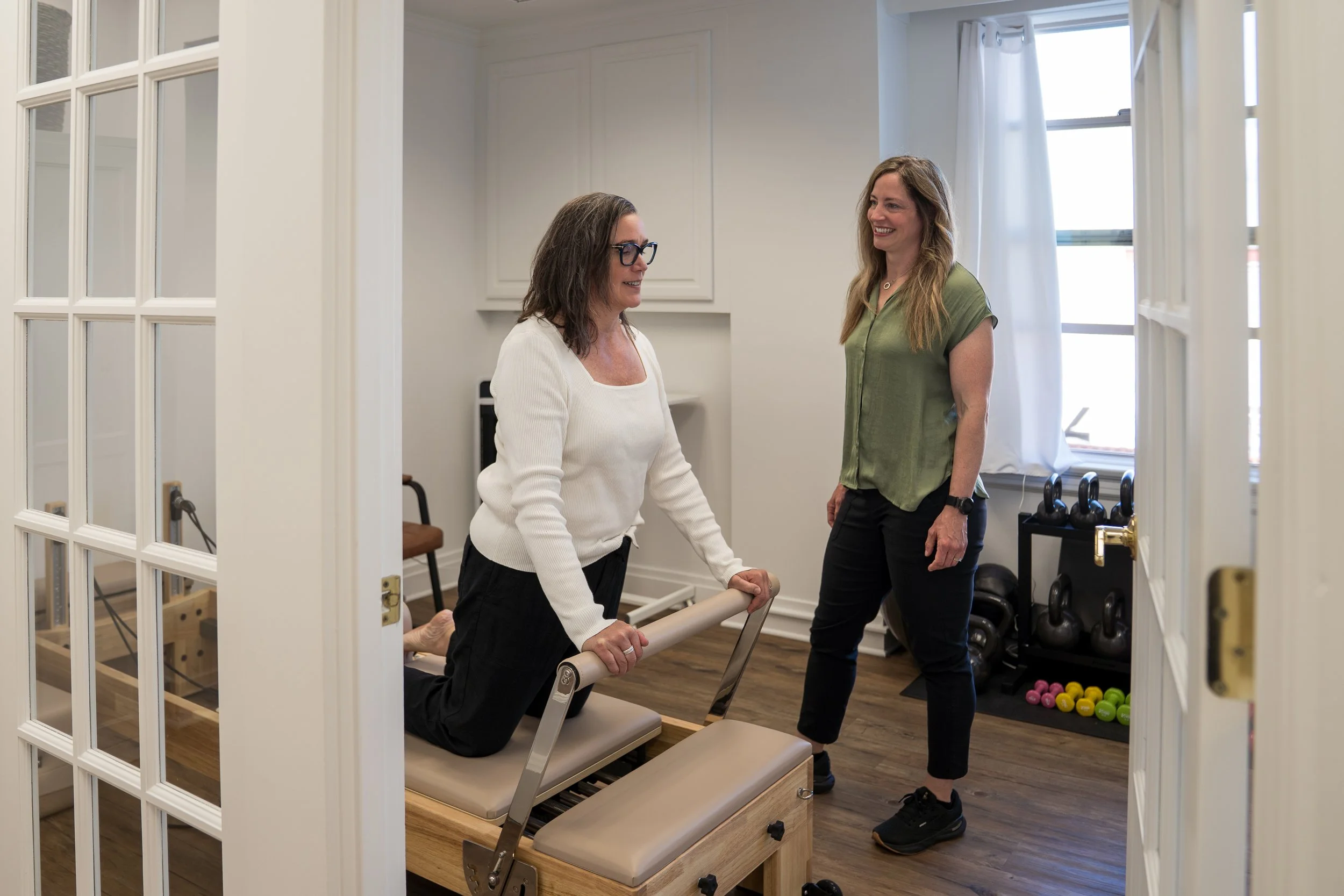 Two women are in a health or fitness setting, one using a Pilates reformer machine, and the other standing nearby, smiling. They are inside a room with large windows, white walls, and athletic equipment like kettlebells and colorful balls.