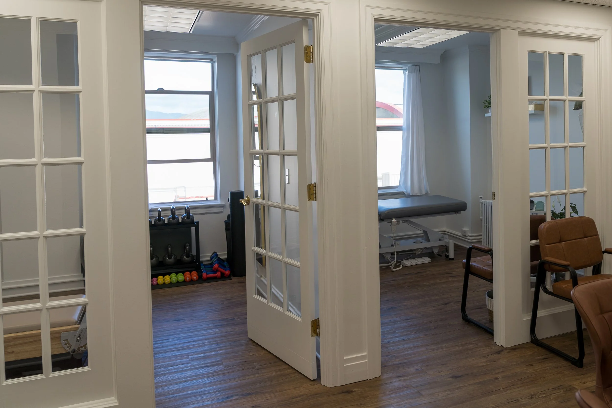A medical room or clinic with exercise and therapy equipment, including dumbbells, kettlebells, and a massage table, separated by glass doors and windows.