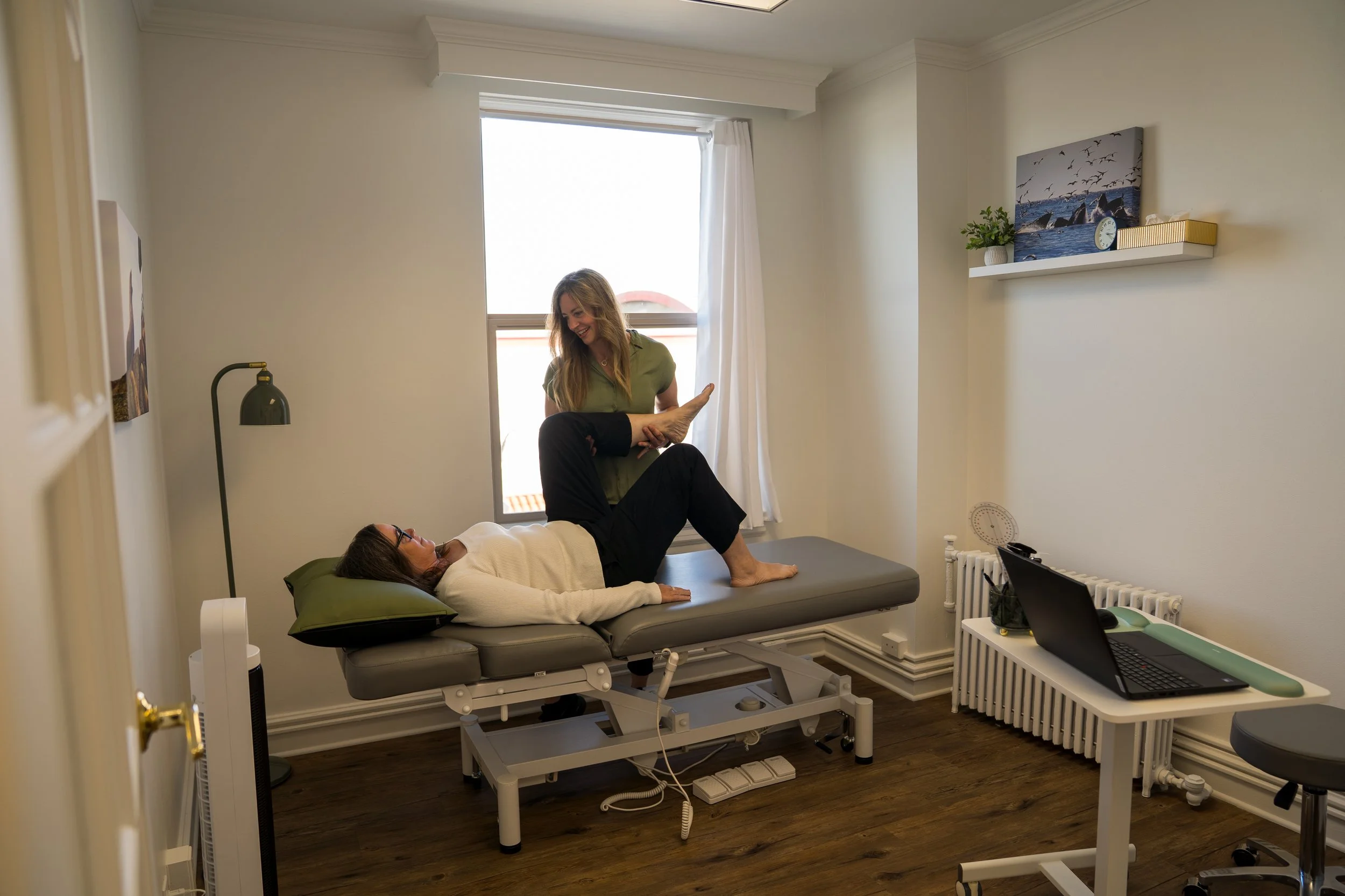 A woman lying on a medical examination table with her legs raised, another woman assisting her, in a medical office with white walls, a window, and a shelf with decorative items.