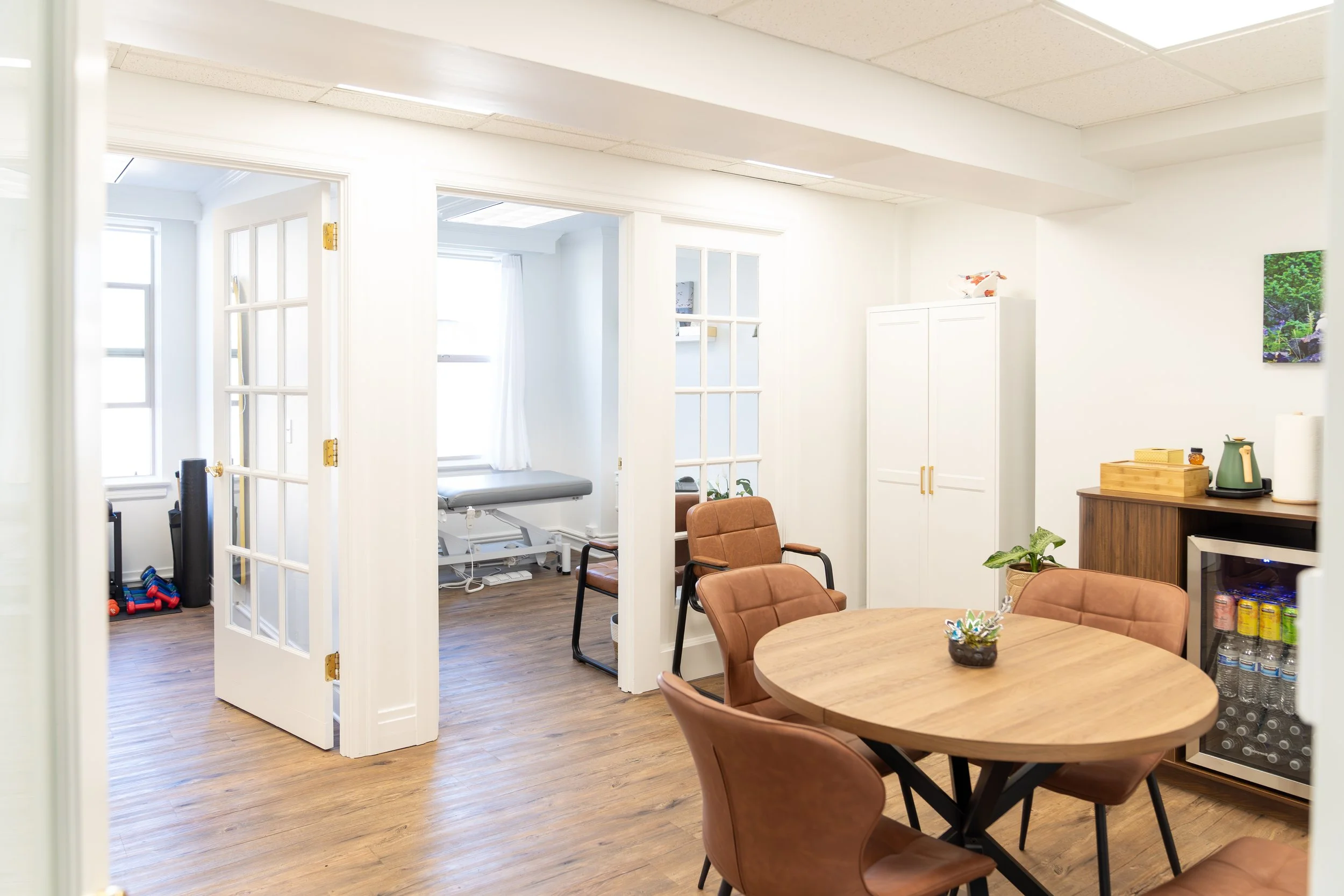 A dining area with a round wooden table and brown chairs, a white cabinet, a small lamp, and a wall with a colorful landscape artwork. In the background, a doorway leads to a room with windows and a chair, and exercise equipment is visible.