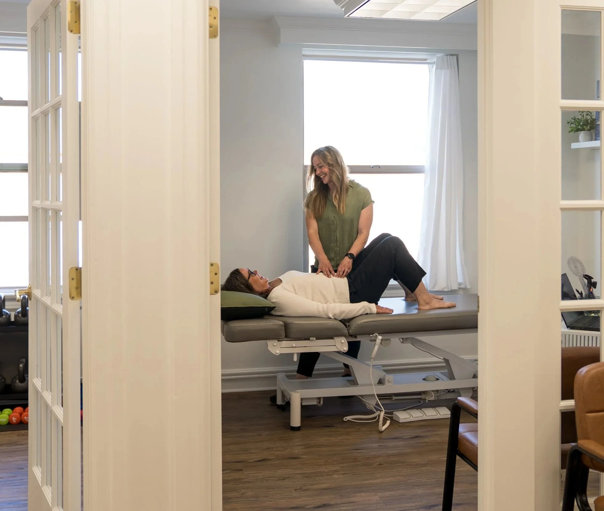 A woman lying on a medical examination table with her legs bent and her head resting on a pillow, while another woman stands beside her with her hands on the patient's stomach, in a bright, airy room with a large window and white curtains.