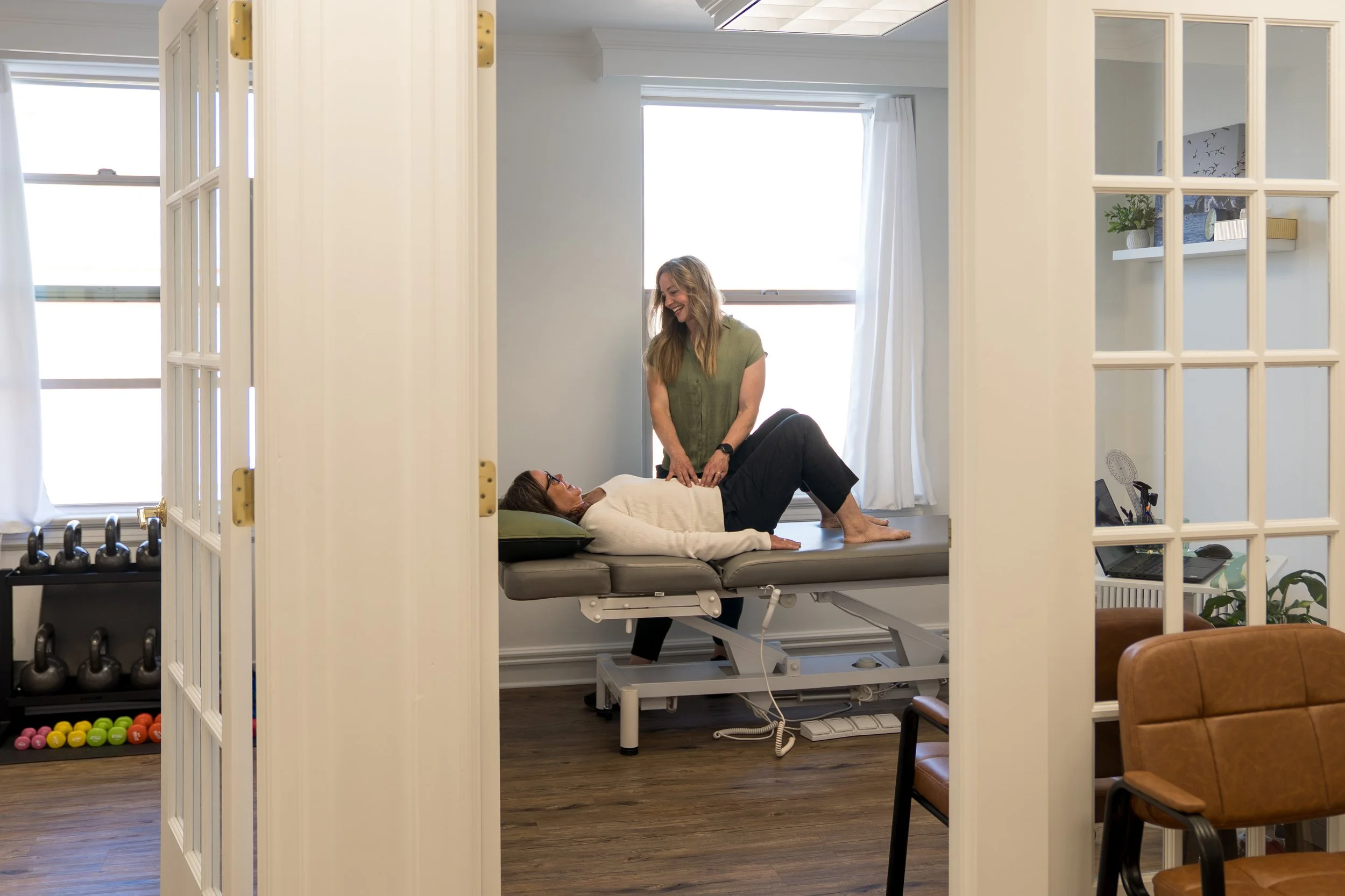 A woman lying on an examination table with her legs bent, wearing glasses and a white sweater, while a woman standing next to her, smiling and touching her abdomen. The room has large windows with white curtains, shelves with potted plants and decor, and gym equipment in the background.
