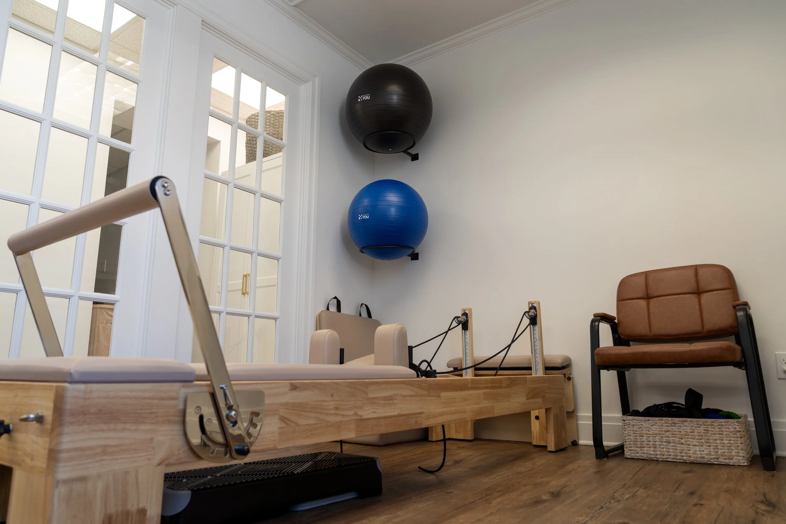 Indoor physical therapy or exercise room with Pilates reformer machine, exercise balls, chair, and resistance bands, with large windows and hardwood flooring.