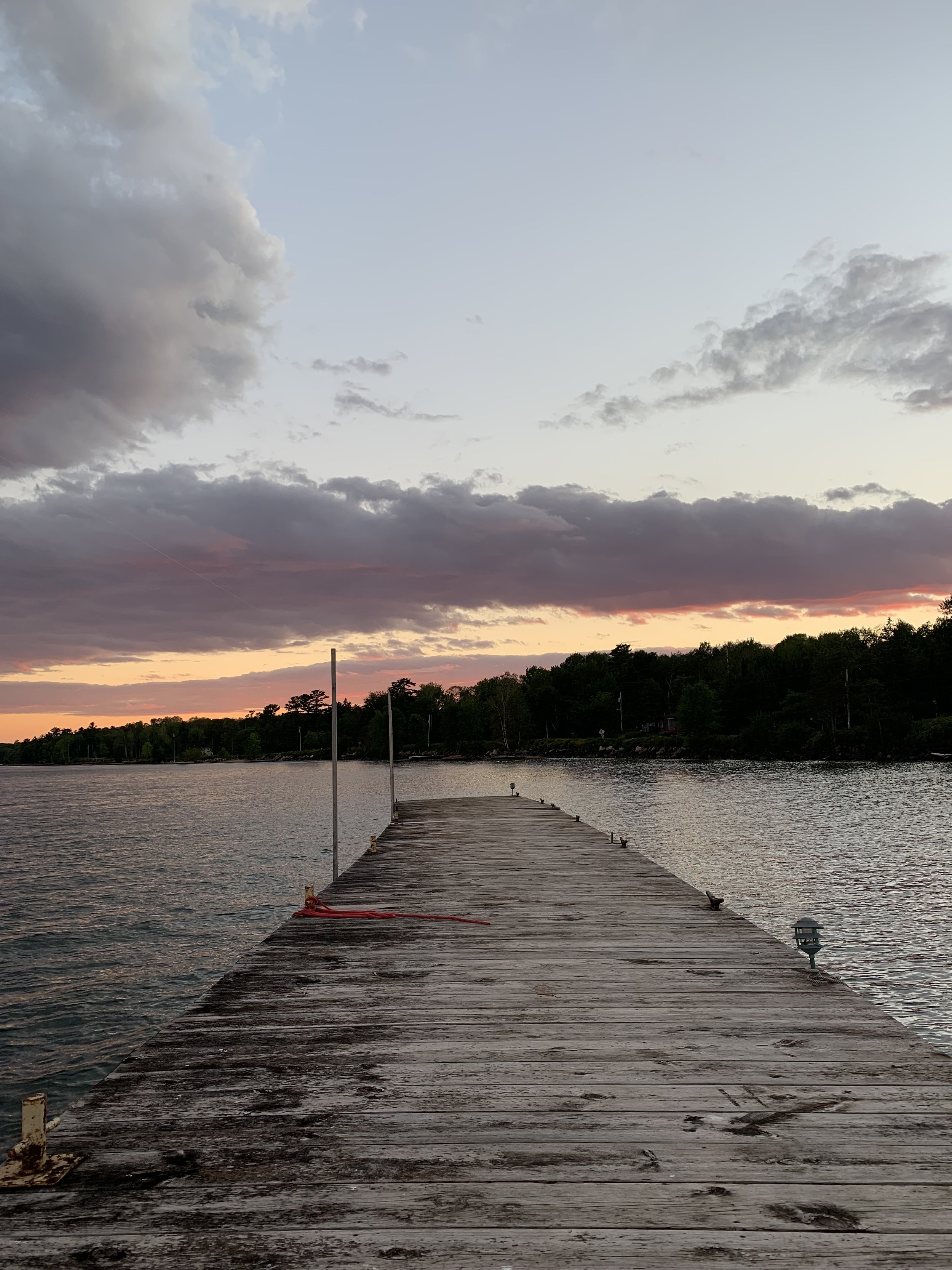 A wooden pier extending into a body of water at sunset, with trees along the shoreline and clouds in the sky.