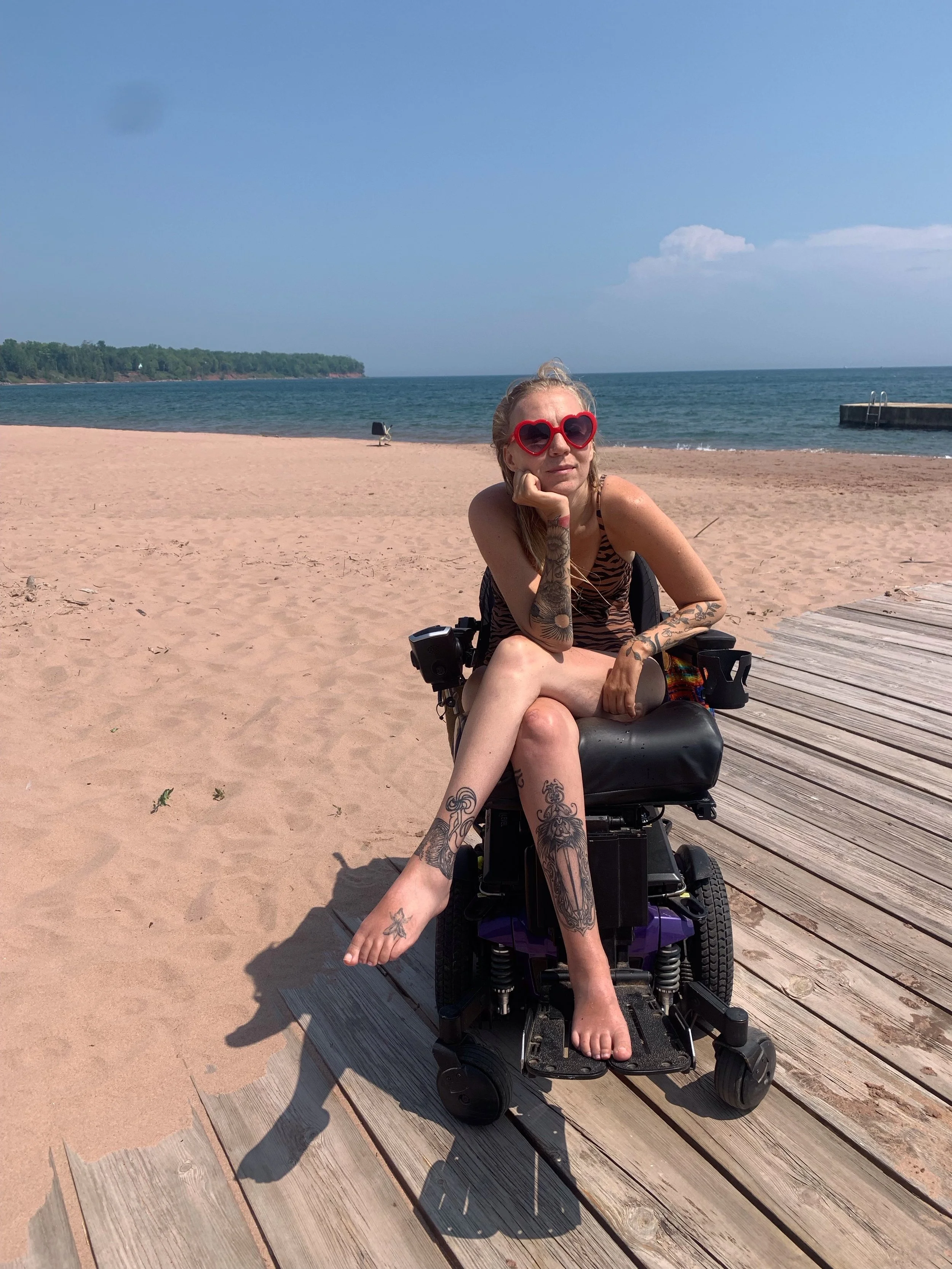 A woman with tattoos, wearing heart-shaped sunglasses, sitting in a power wheelchair on a sandy beach with the lake and a blue sky in the background.