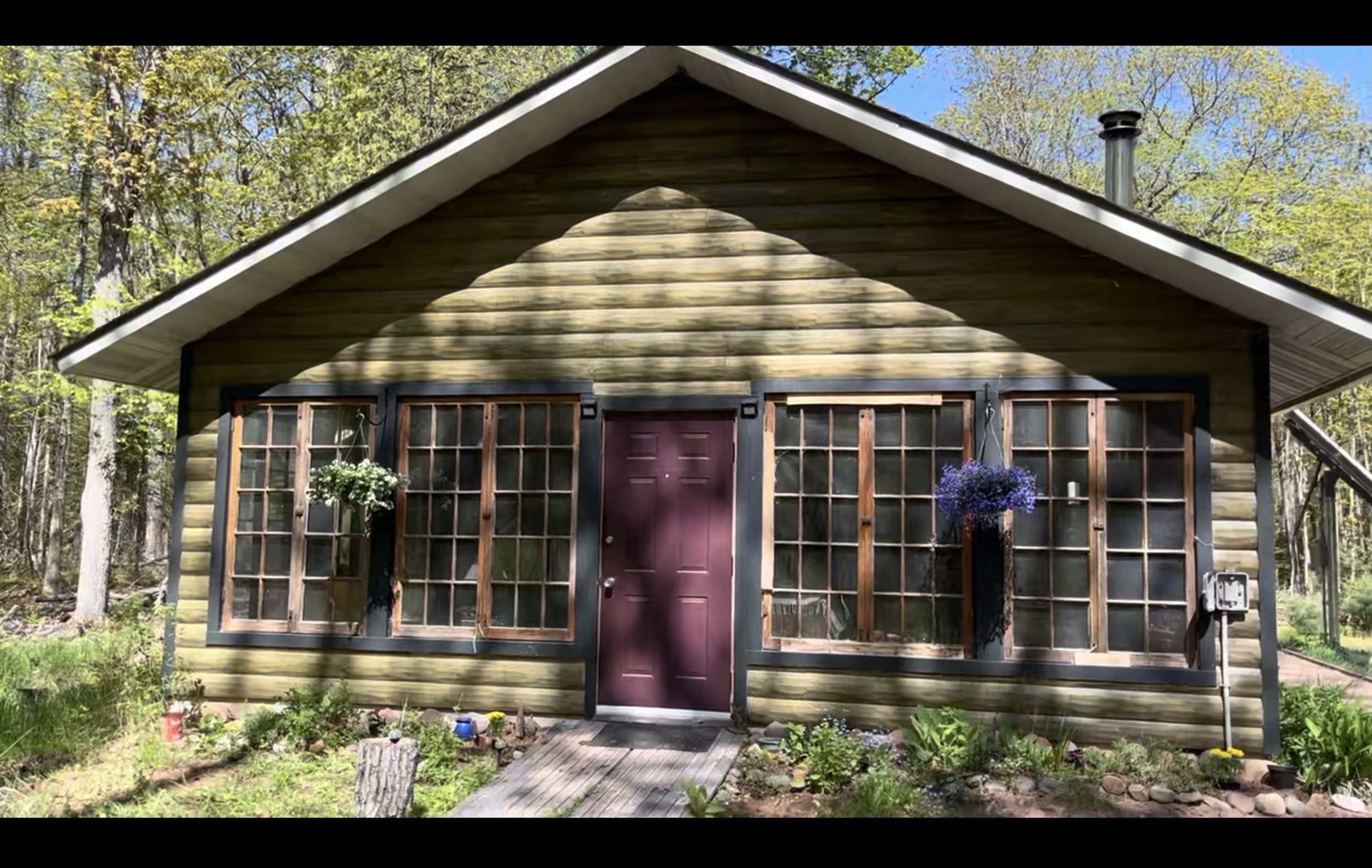 A small wheelchair-accessible house with a wooden exterior, pine frame windows, a purple front door, and a sloped roof,  and off-grid living surrounded by the forest.