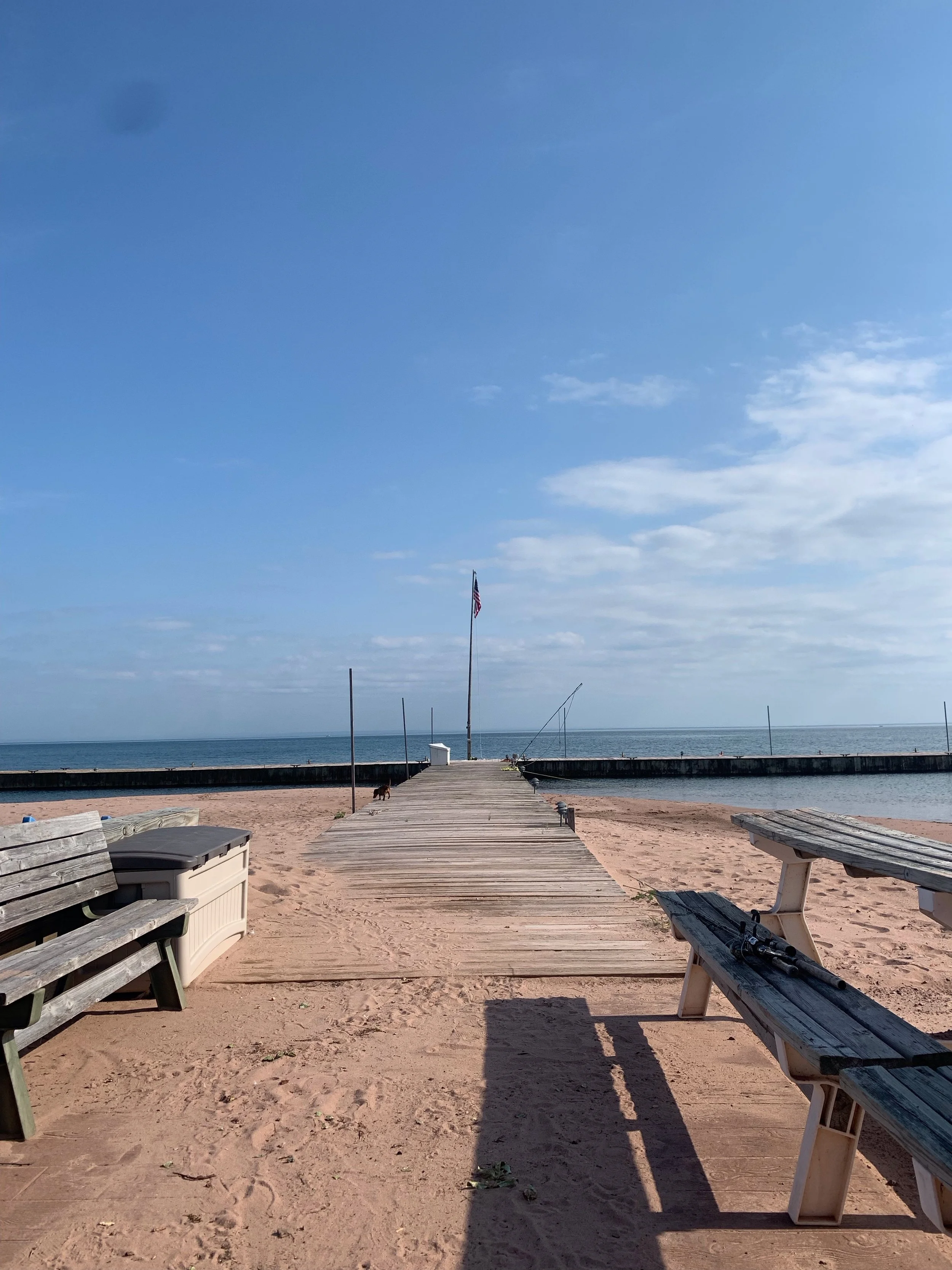 A sandy beach with an accessible wooden dock leading out onto the water, flanked by benches and a flagpole, under a partly cloudy sky.