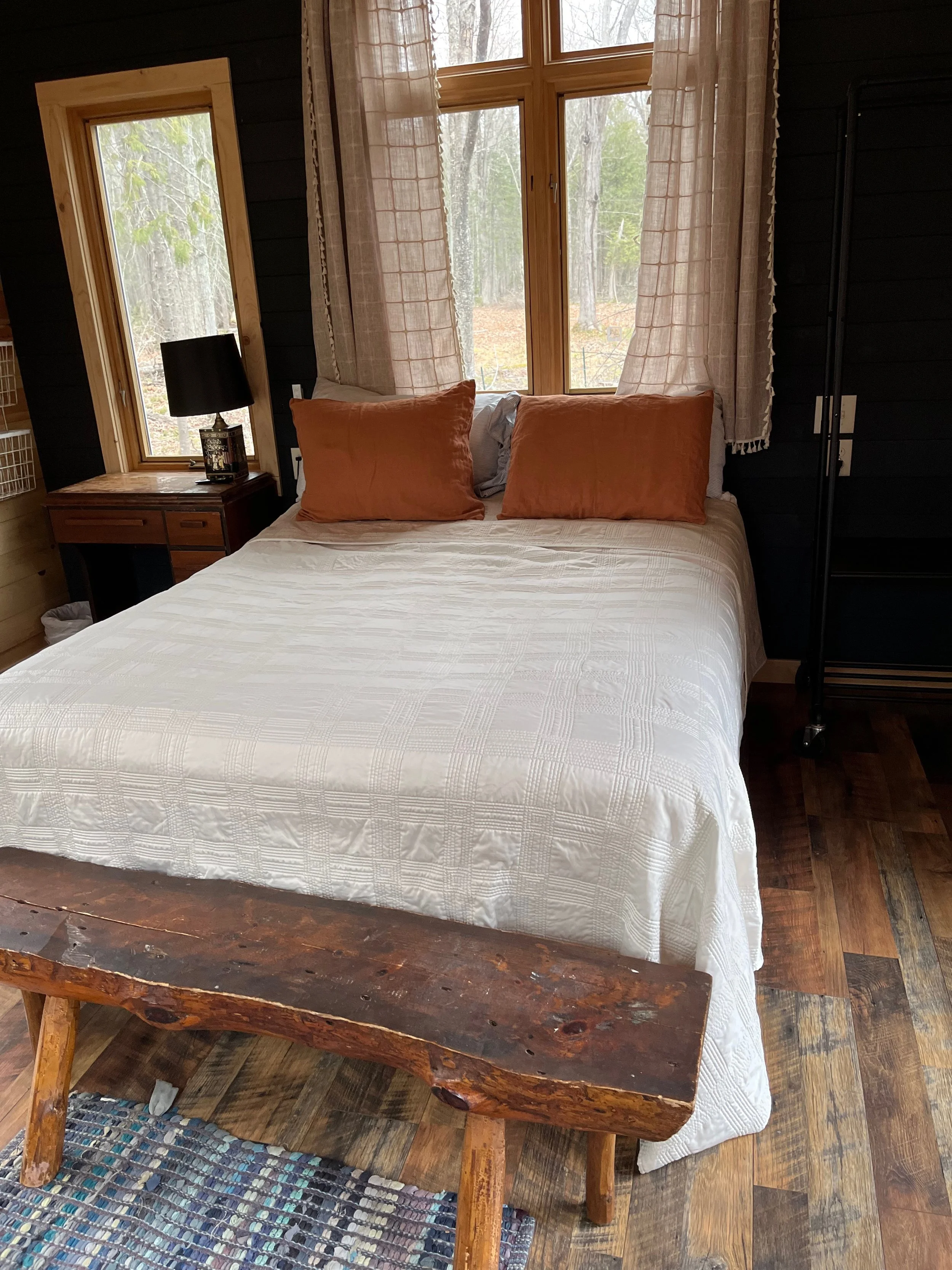View of a bedroom with large windows, a white bed with orange pillows, a wooden bedside table with a black lamp, curtains, and a rustic wooden bench at the foot of the bed. Outside trees are visible through the windows.