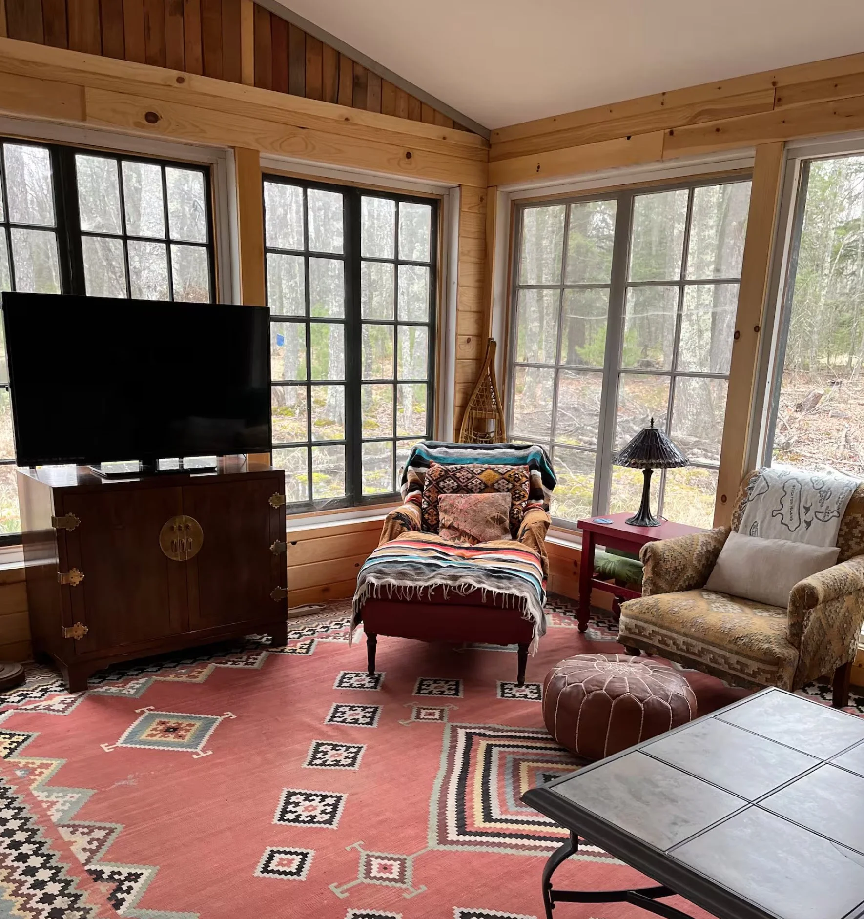 Living room with large windows showing a wooded outdoor scene, decorated with patterned rug, vintage armchair with blanket, wooden furniture, and lamps.
