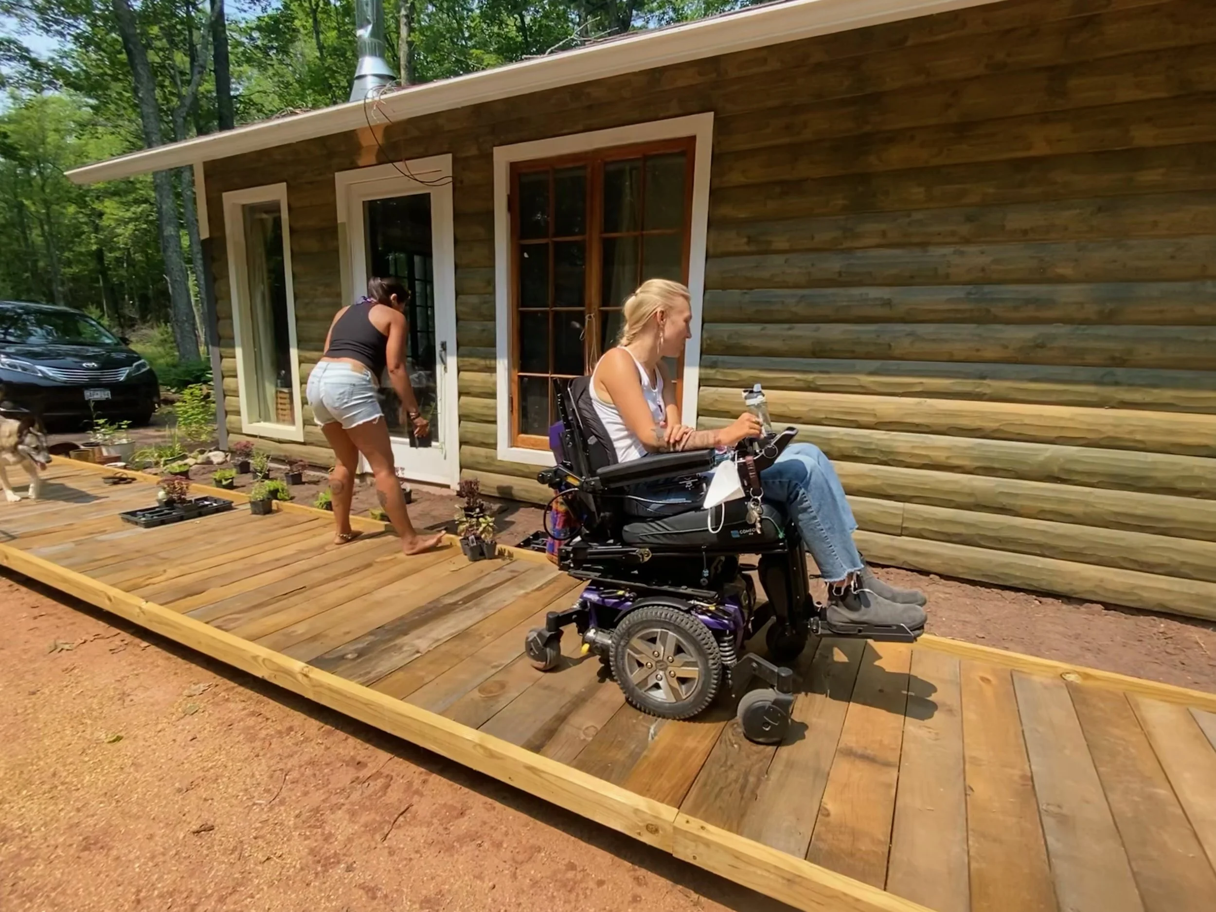 Women gardening outside on an accessible deck, surrounded by trees, one woman is sitting in a power wheelchair, another woman is planting nearby.