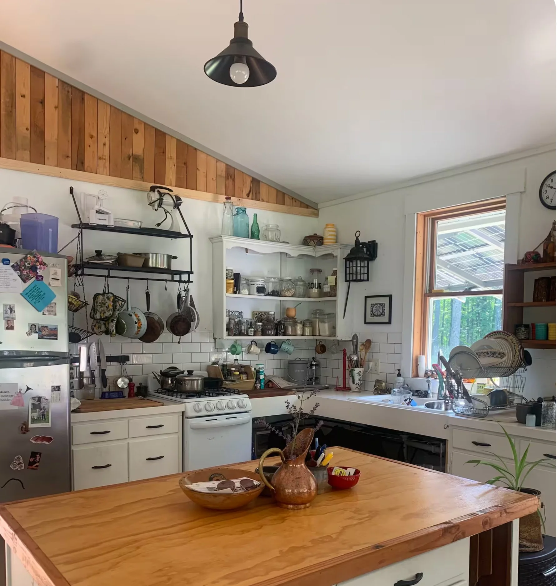 A kitchen with a wooden countertop, white cabinets, and open shelves. There are jars, bottles, and kitchen utensils on the shelves. A window with a view of solar panels on the roof. A ceiling light fixture hangs from the ceiling. The fridge is covered with magnets and photos.