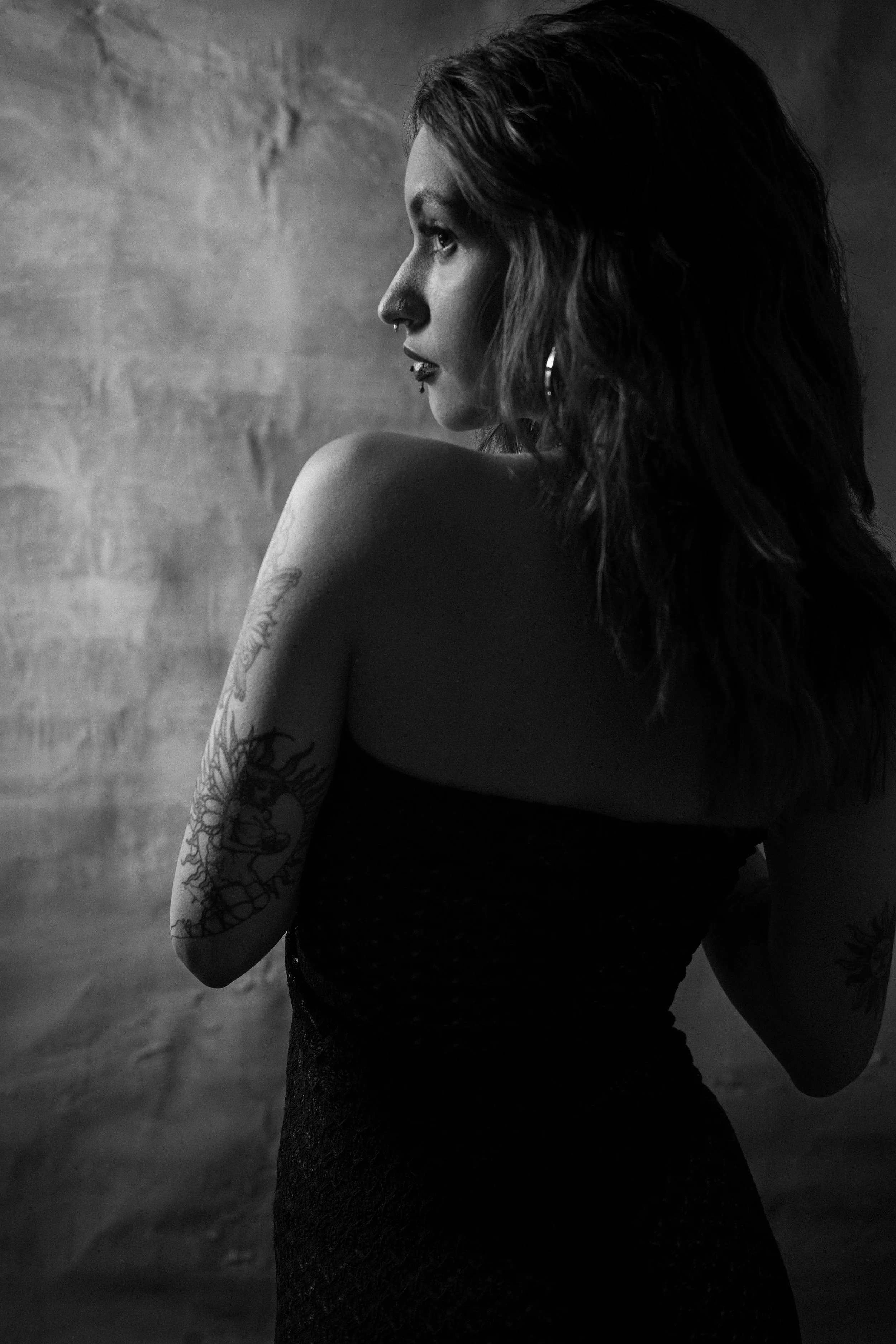 Black and white portrait of a woman with wavy hair, tattoos on her arm, wearing earrings and a strapless dress, standing against a textured wall.