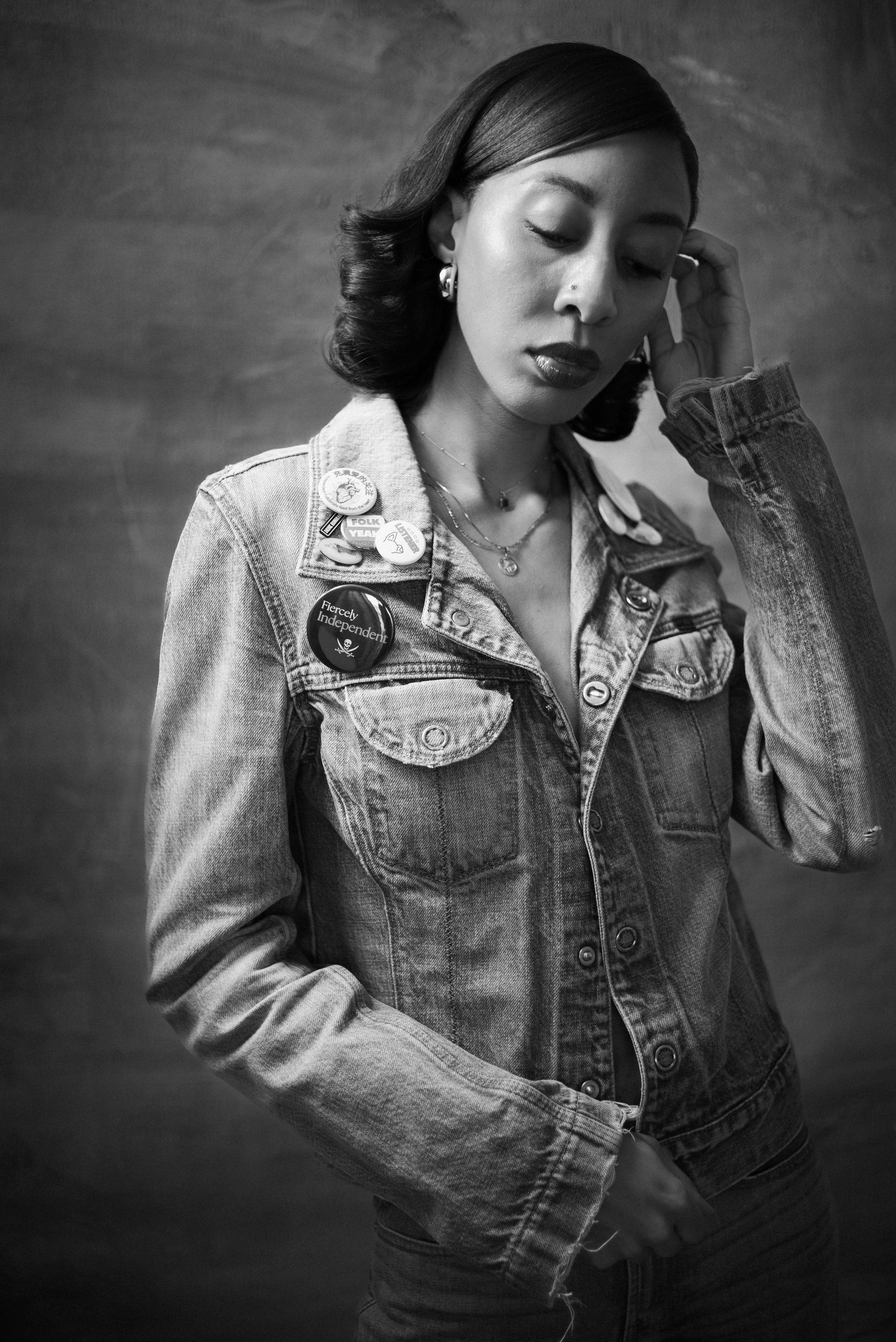 Black and white portrait of a woman wearing a denim jacket with buttons and pins, looking down with her hand near her face.
