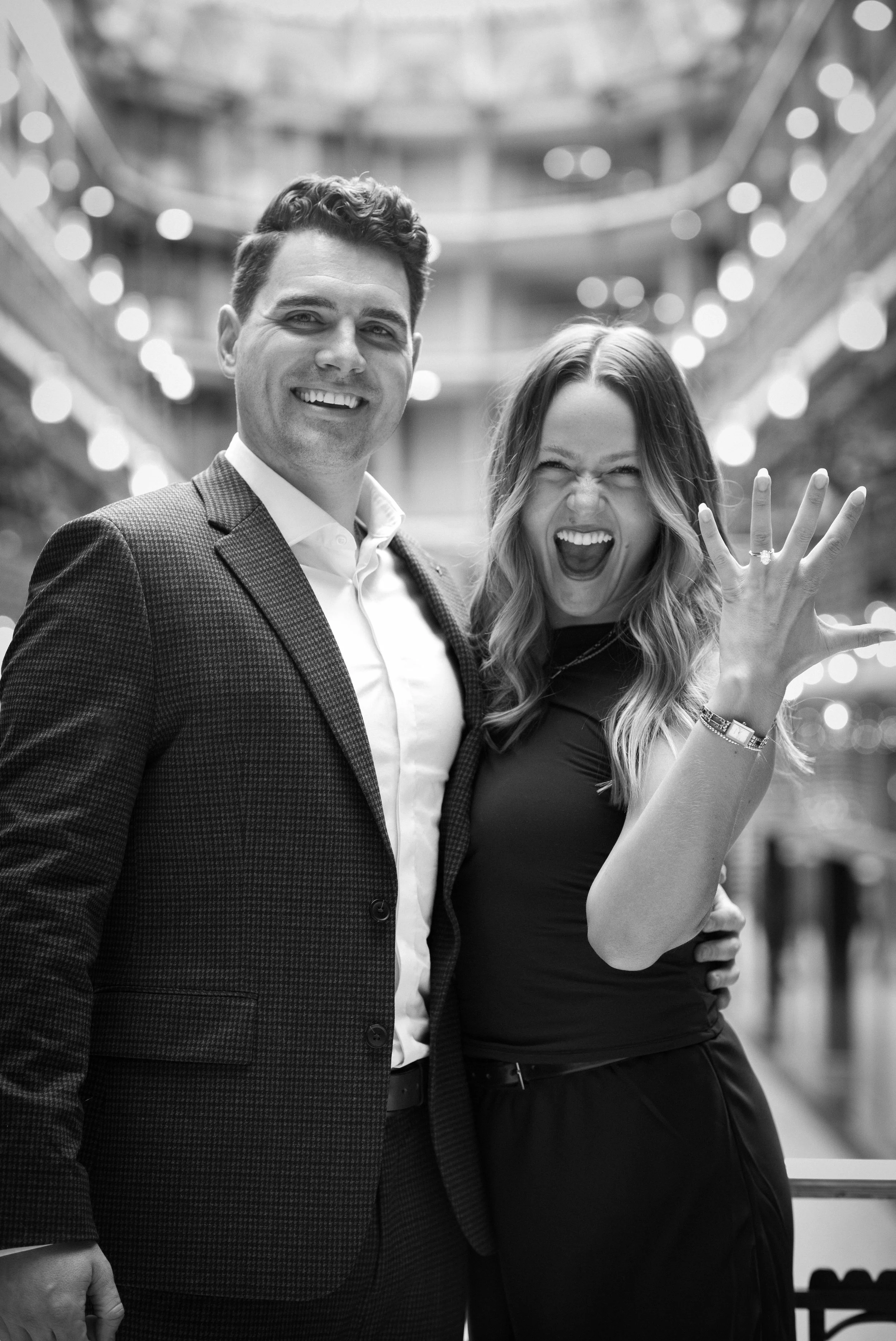 A smiling man and a woman celebrating an engagement, with the woman showing off a ring on her finger, in an indoor setting with blurred lights in the background.