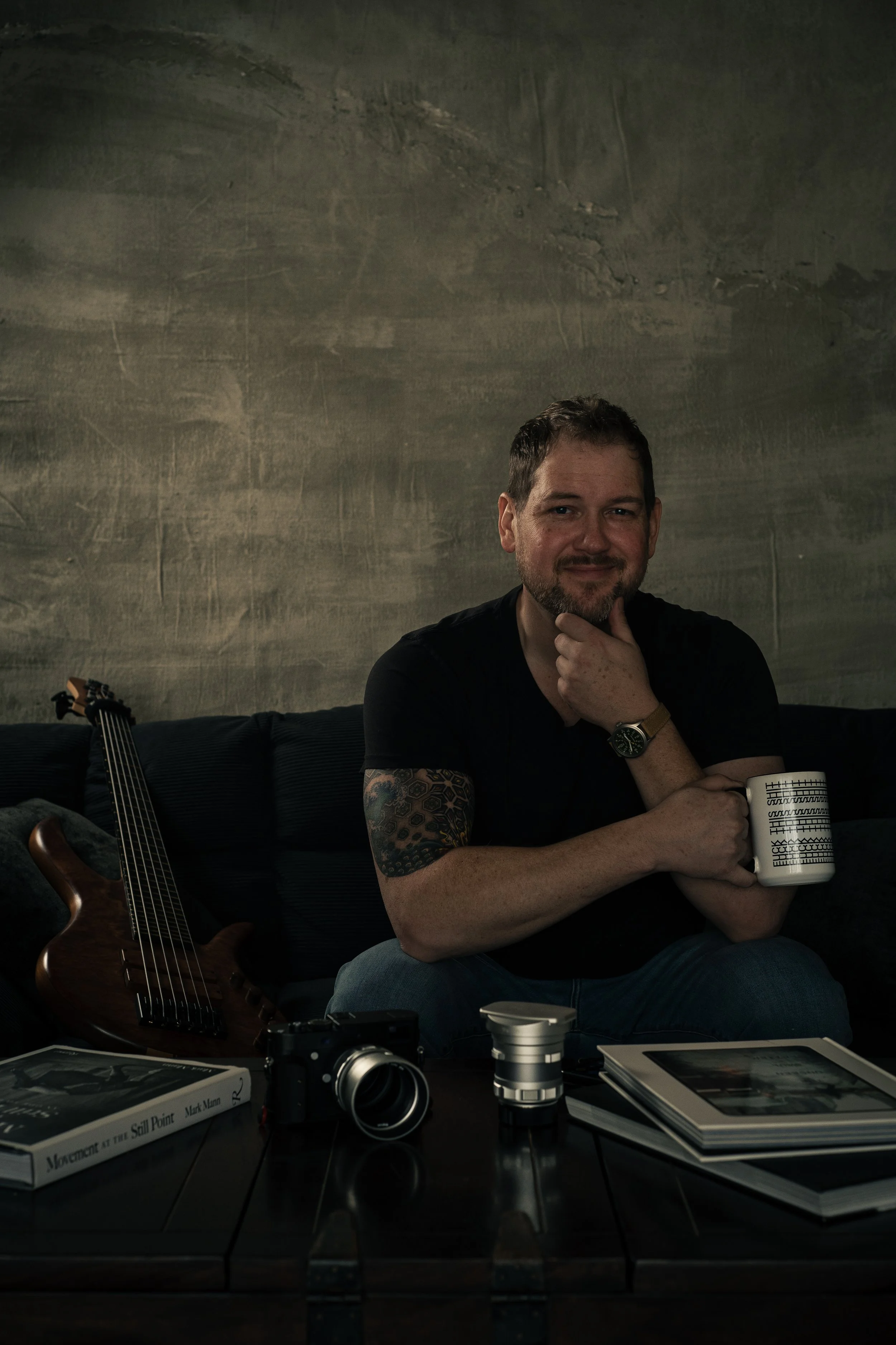 A man sitting on a couch holding a white coffee mug with the word "SHAT" printed repeatedly, a guitar resting beside him, and a table with a camera, lenses, and books in front of him, against a textured wall background.
