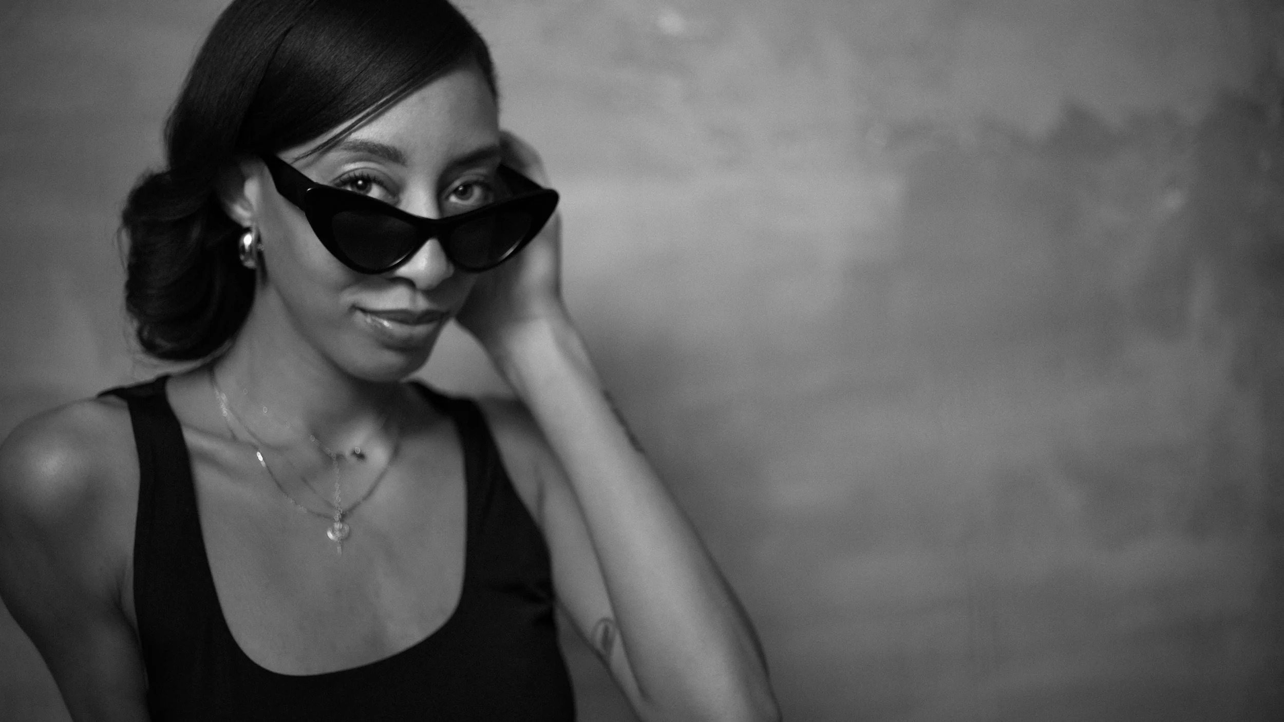 A woman with dark hair, wearing sunglasses and jewelry, looking at the camera in a black and white photo.