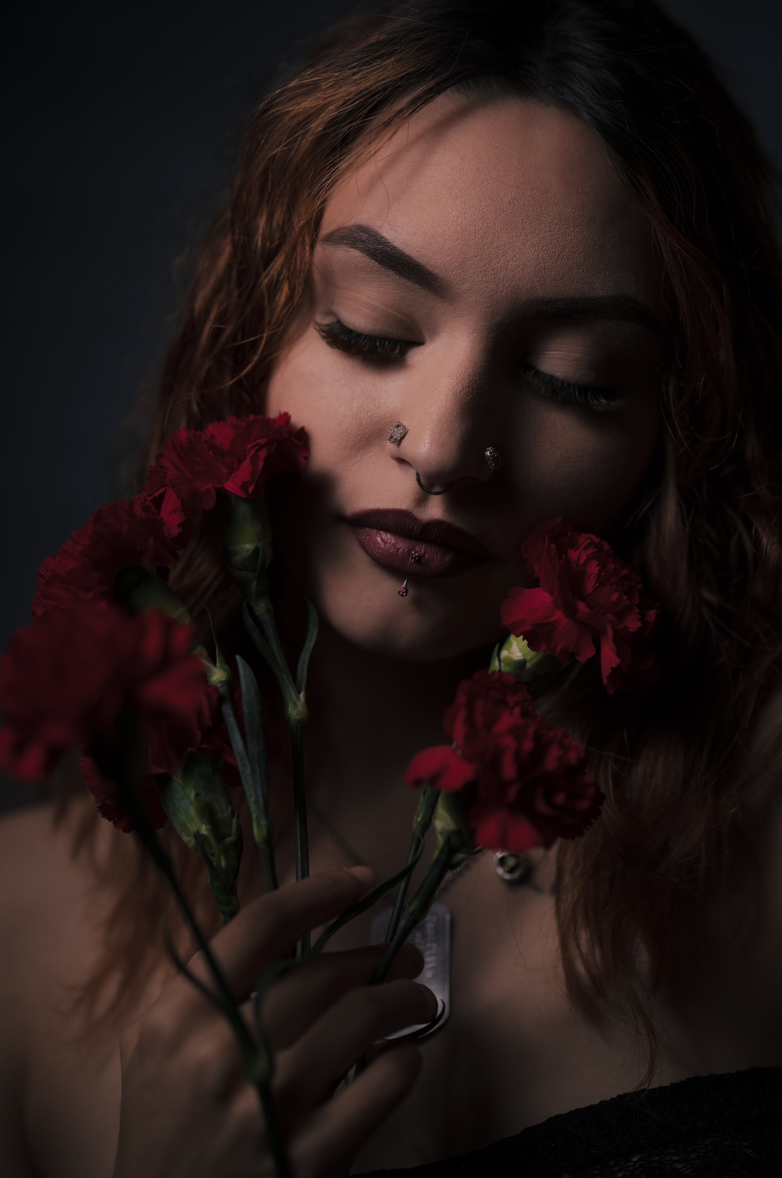 A woman with closed eyes and cranial piercings, holding red carnations, dark background.