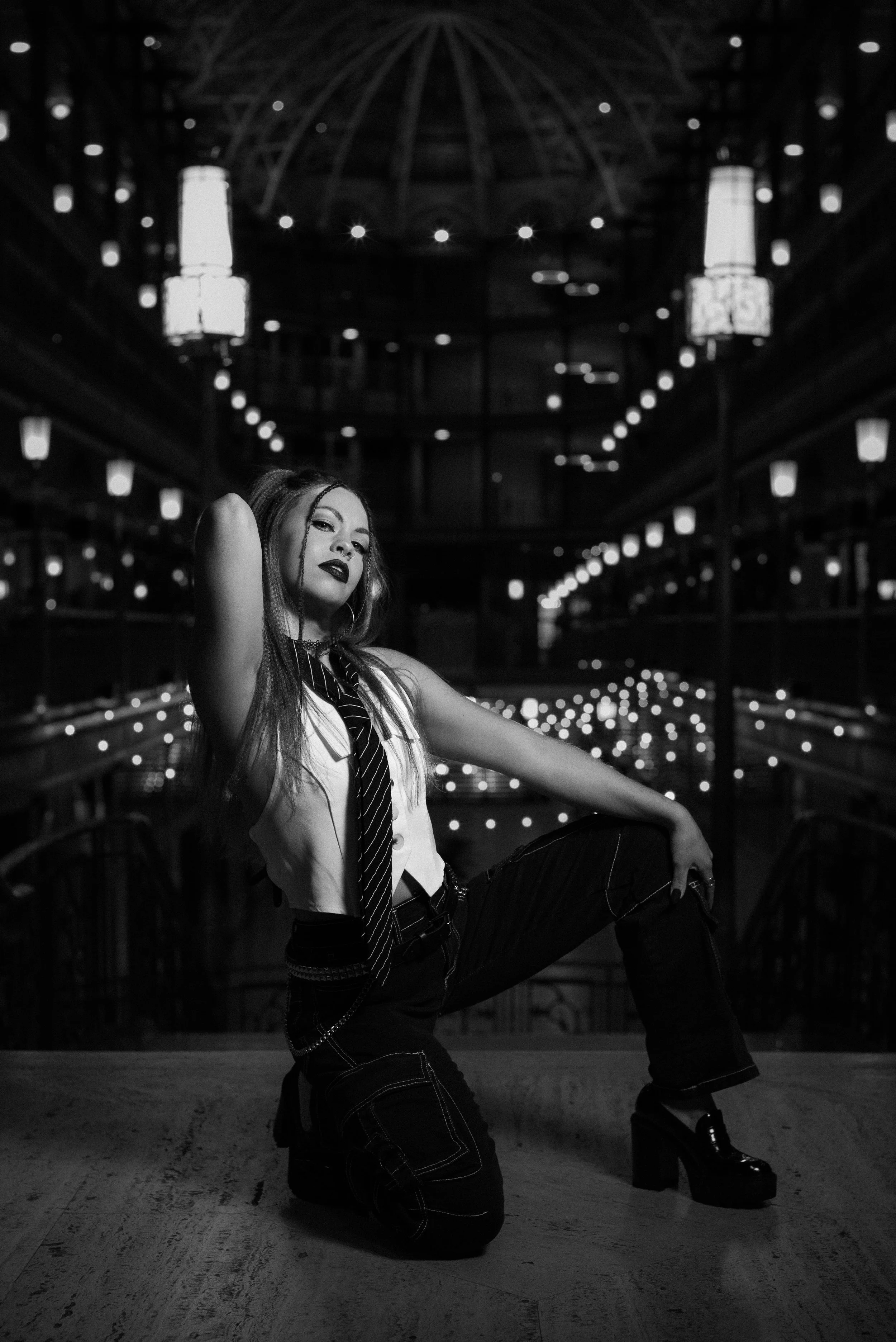 A young woman with long hair and dark lipstick posing confidently in a dimly lit, elegant interior with hanging lamps and reflective surfaces, dressed in a sleeveless shirt, tie, ripped jeans, and high heels.