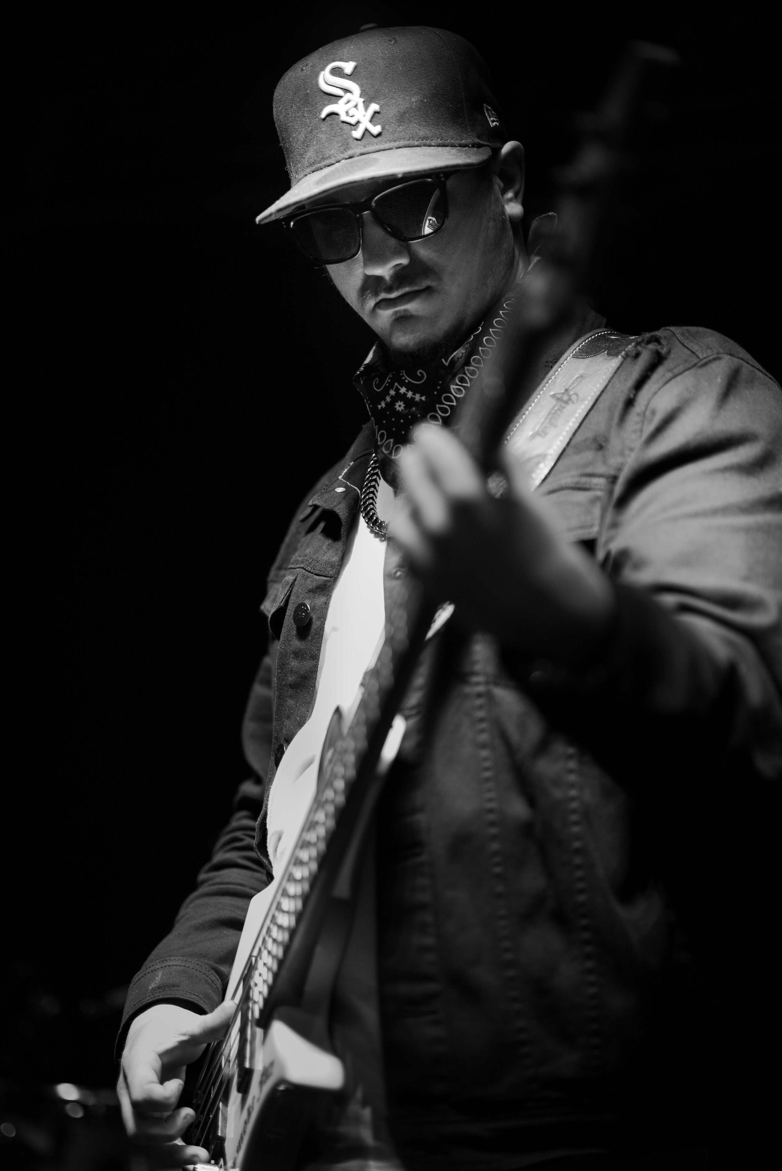 A man wearing sunglasses, a cap with a sport logo, and a bandana around his neck plays an electric guitar in a dark setting.
