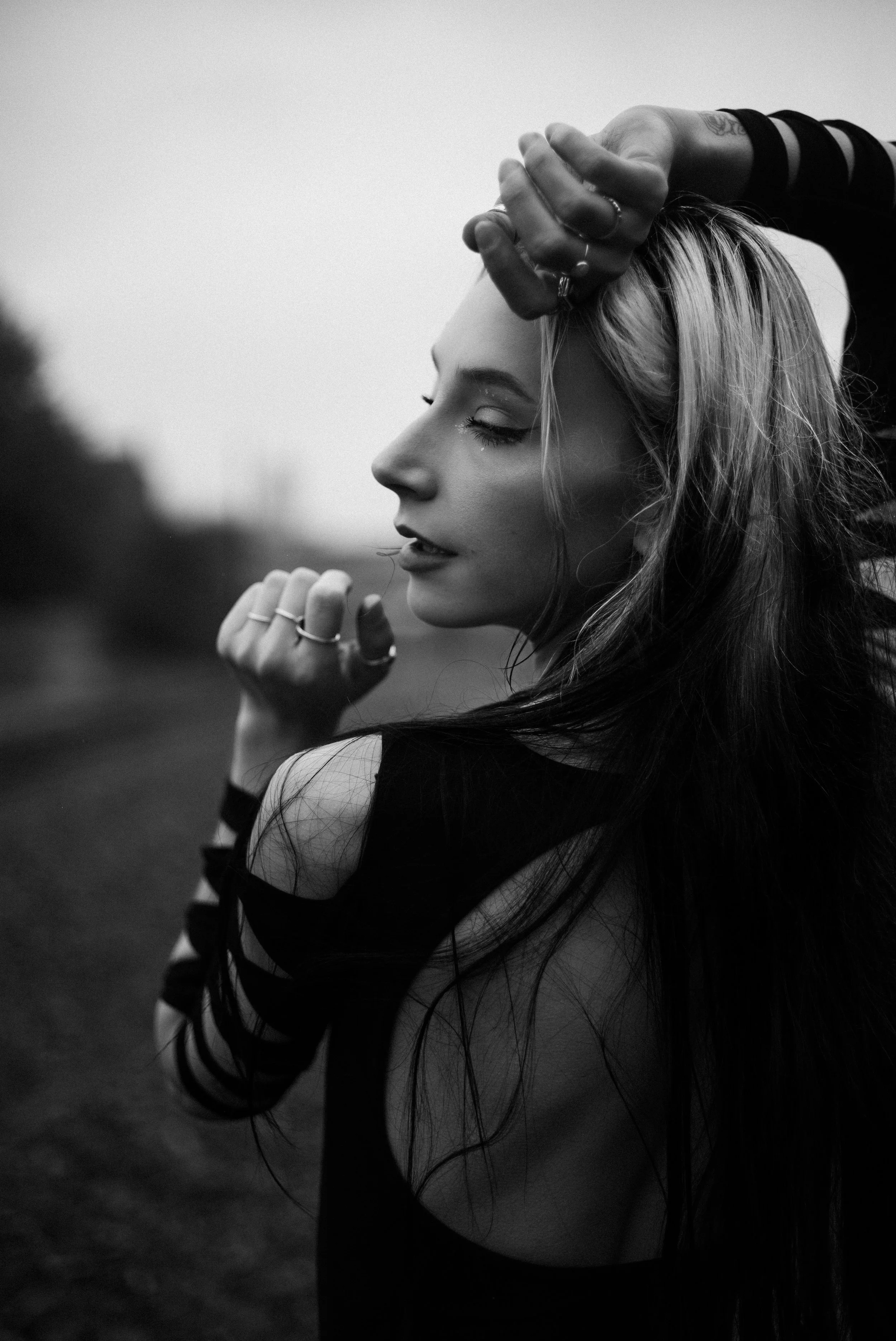 A black and white photo of a woman with long hair, eyes closed, with her hand on her forehead and her other hand near her chin, standing outdoors.
