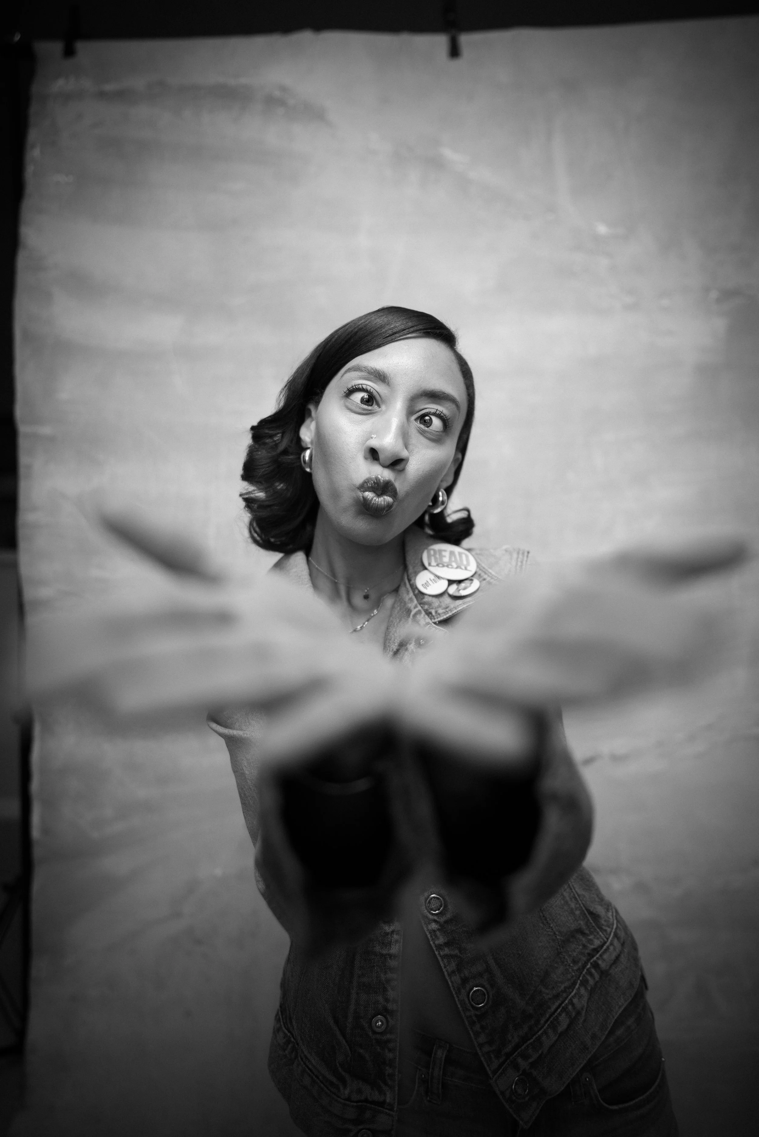A woman taking a selfie with a funny facial expression, puckering her lips, wearing a denim jacket with buttons and pins, standing in front of a plain wall.