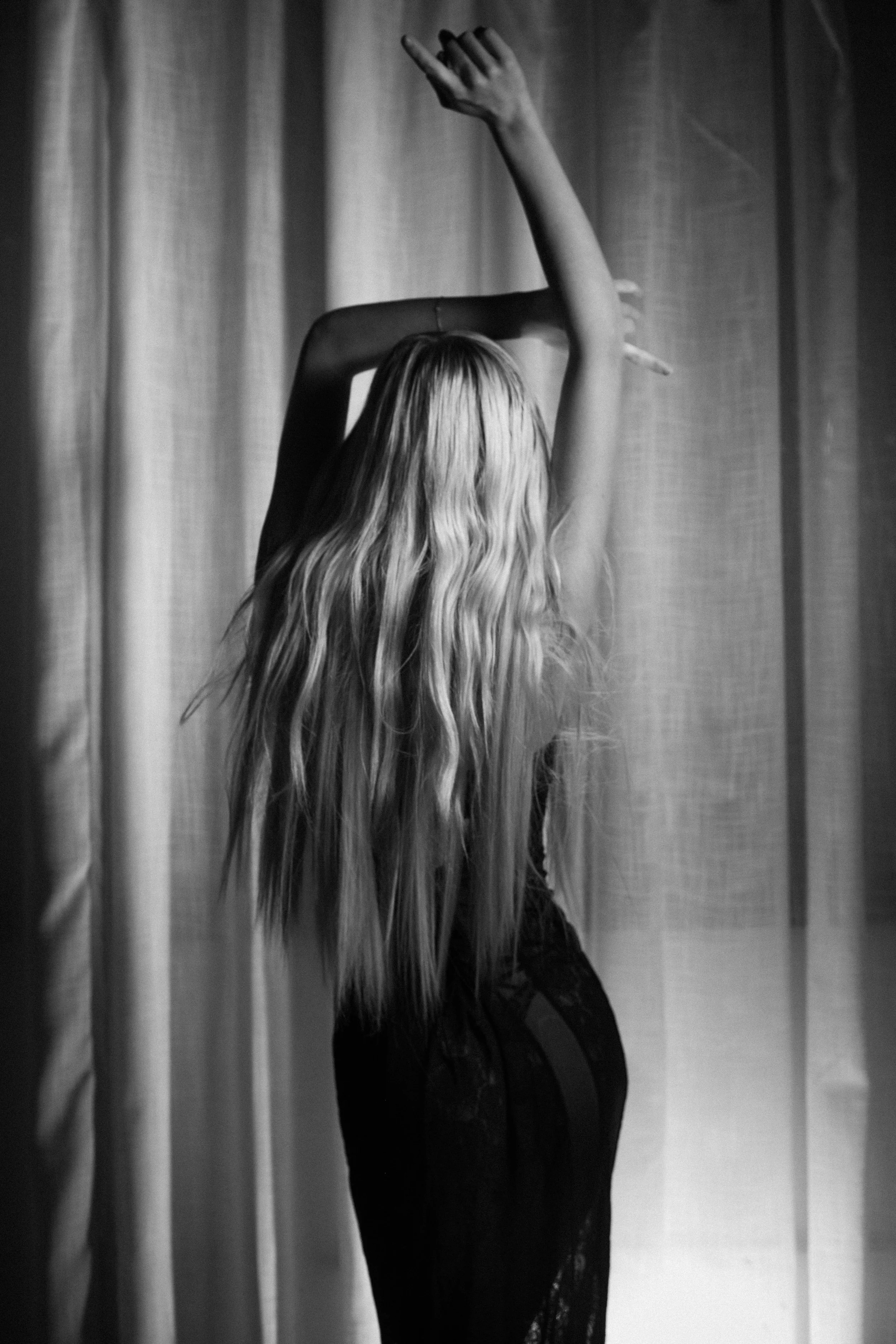 Black and white photo of a woman with long wavy hair, with only one arm visible, raised above her head, against a curtain background.