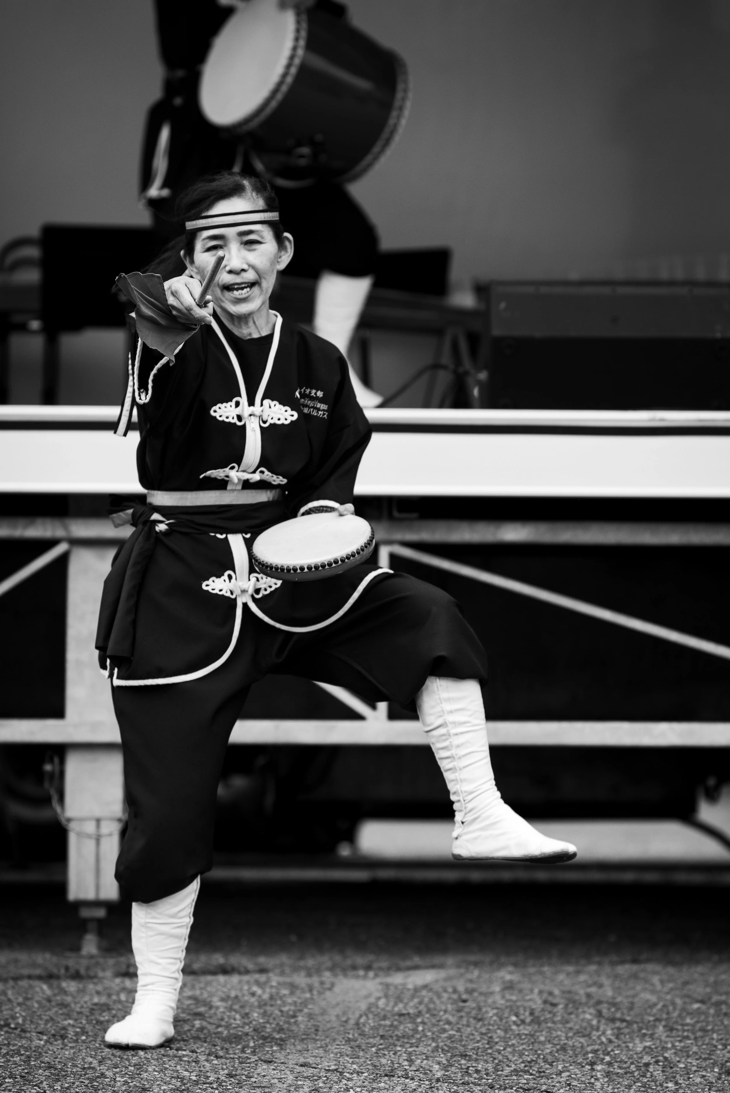 A woman in traditional Japanese attire performs a dance on stage, holding a small drum and a stick, with her leg raised and a joyful expression.