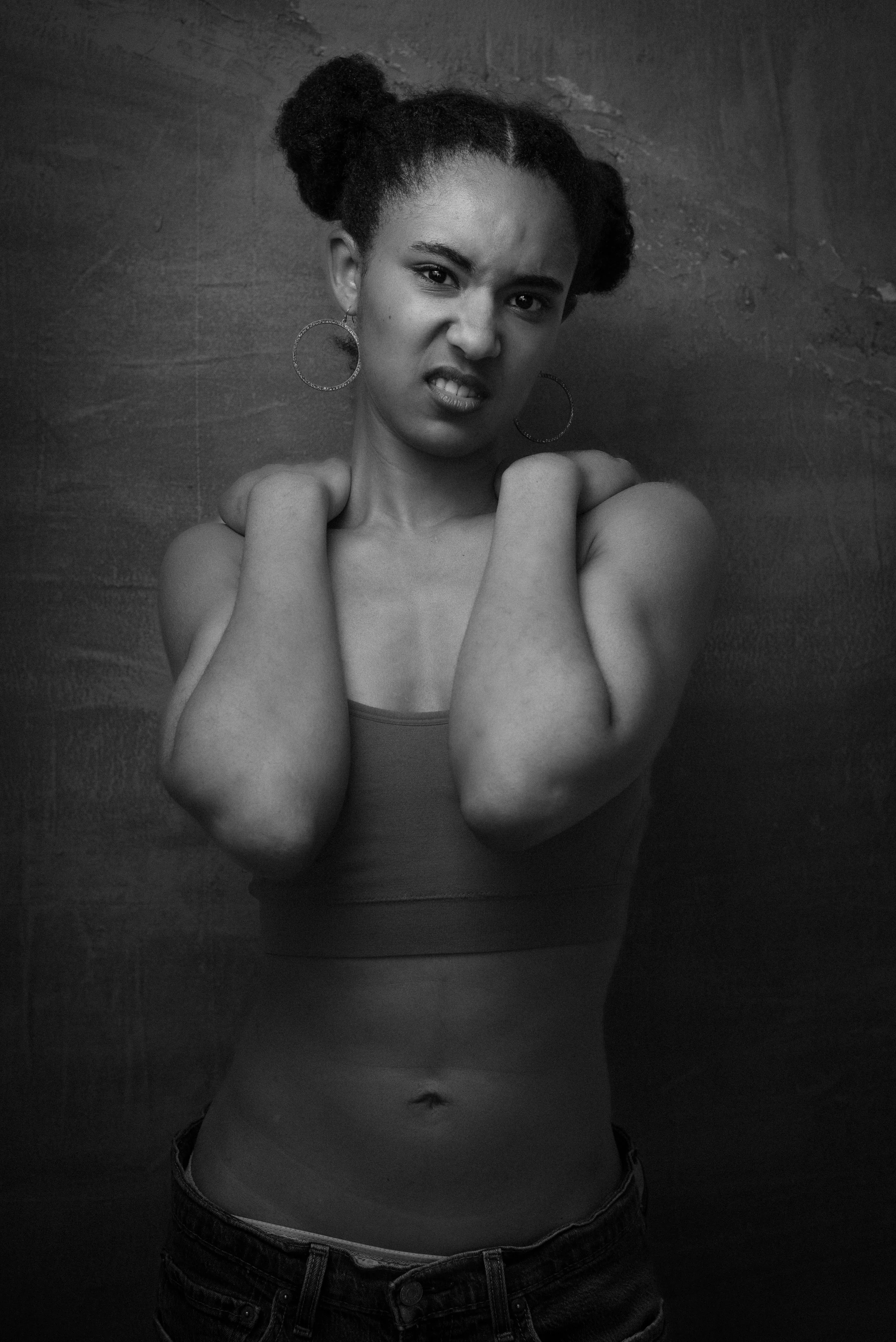 Black and white photo of a woman with her hair styled in two puff ponytails, wearing large hoop earrings and a sleeveless top, standing against a textured wall, making a facial expression with her teeth showing and eyebrows furrowed, with her hands r