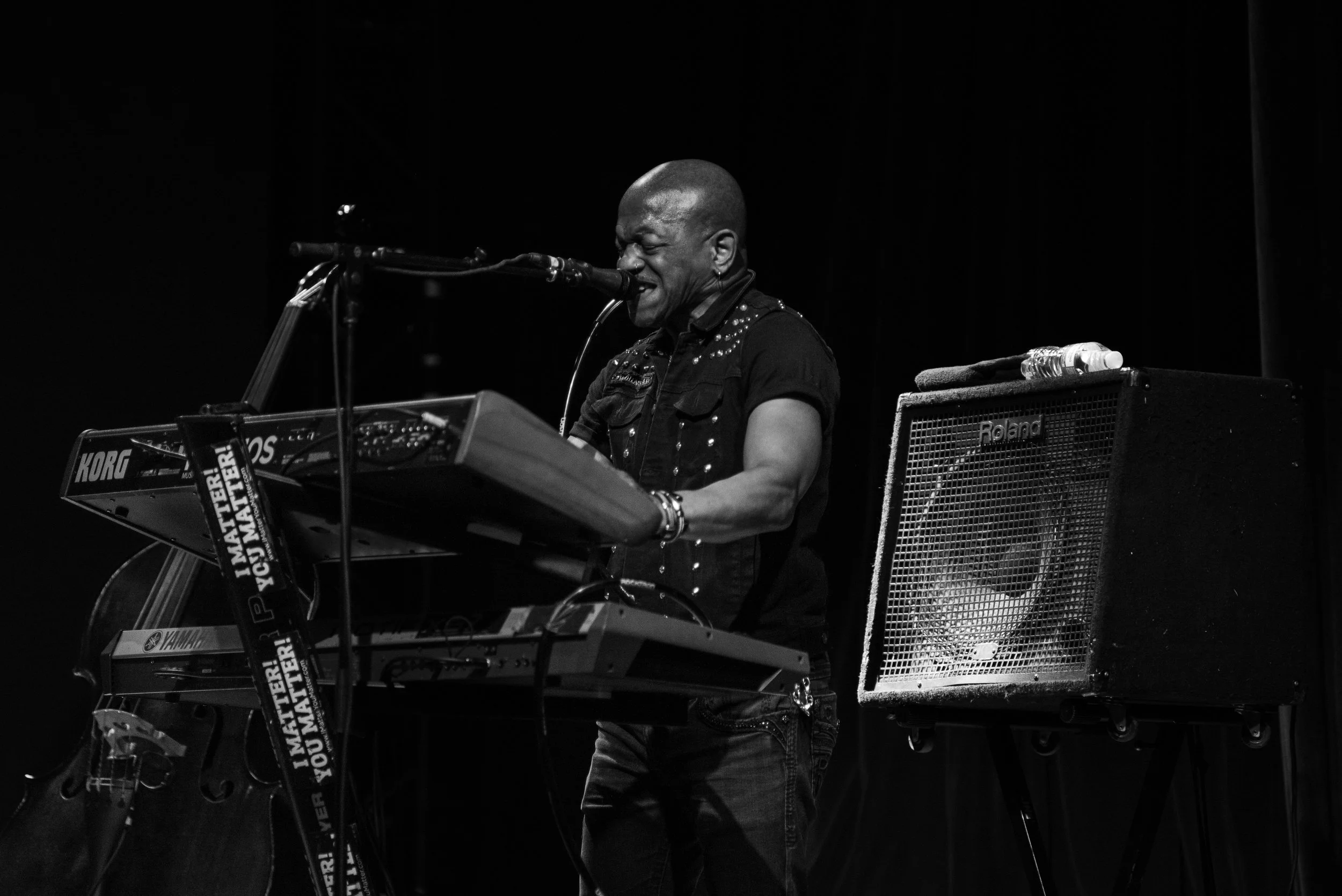A musician performs on stage with a keyboard and microphone, wearing a black shirt with studs, next to a Roland amplifier in a dark setting.