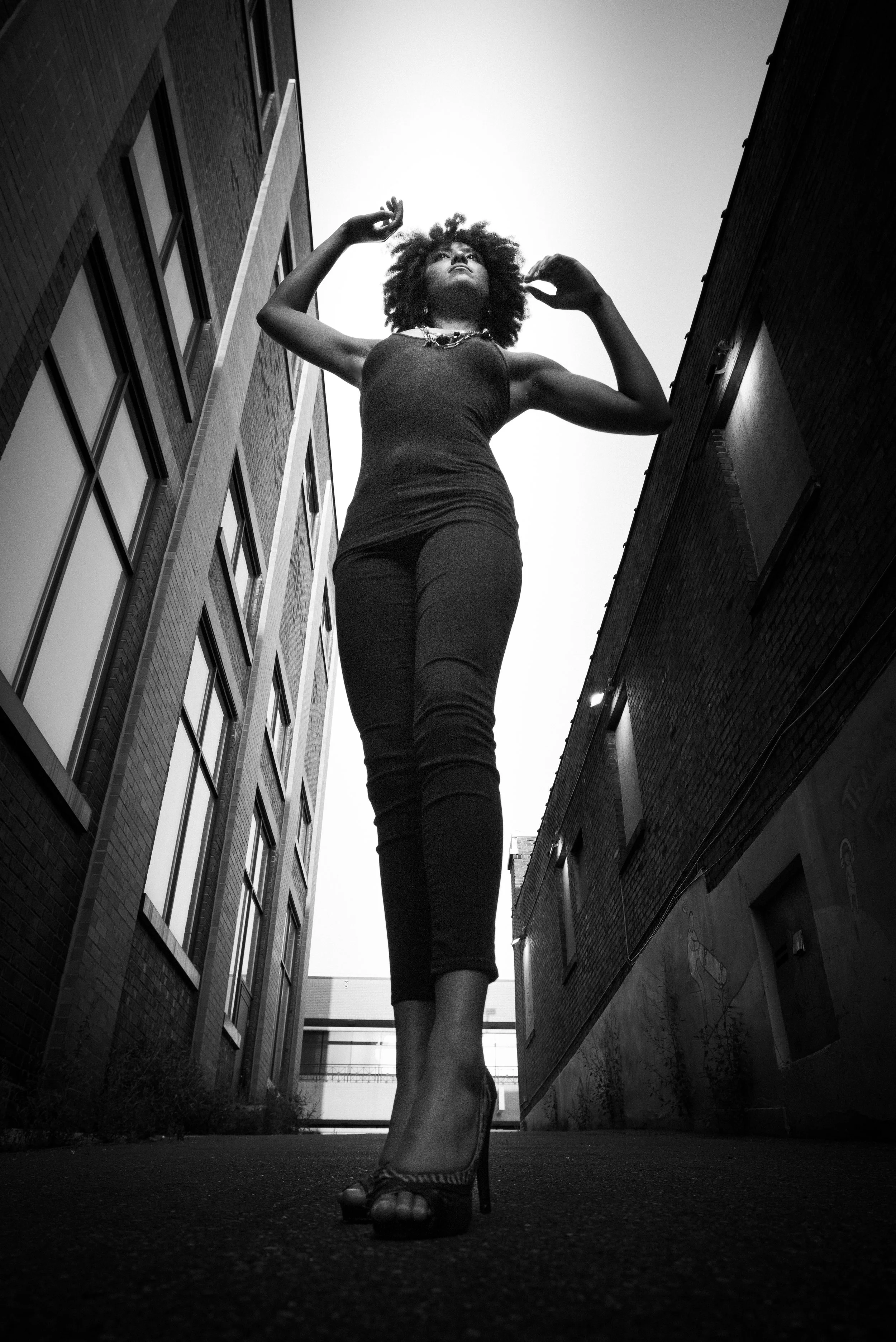 A woman with curly hair wearing high heels, a sleeveless top, and pants, walking between tall buildings in an alley taken from a low-angle shot in black and white.
