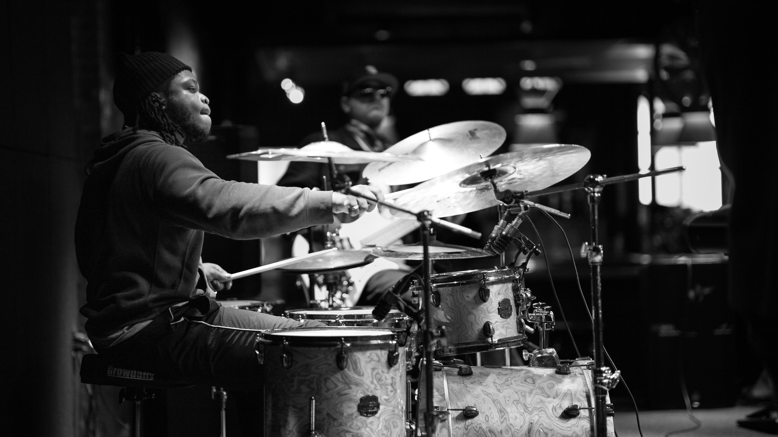 Black and white photo of a drummer playing a drum set on stage, with another musician in the background.