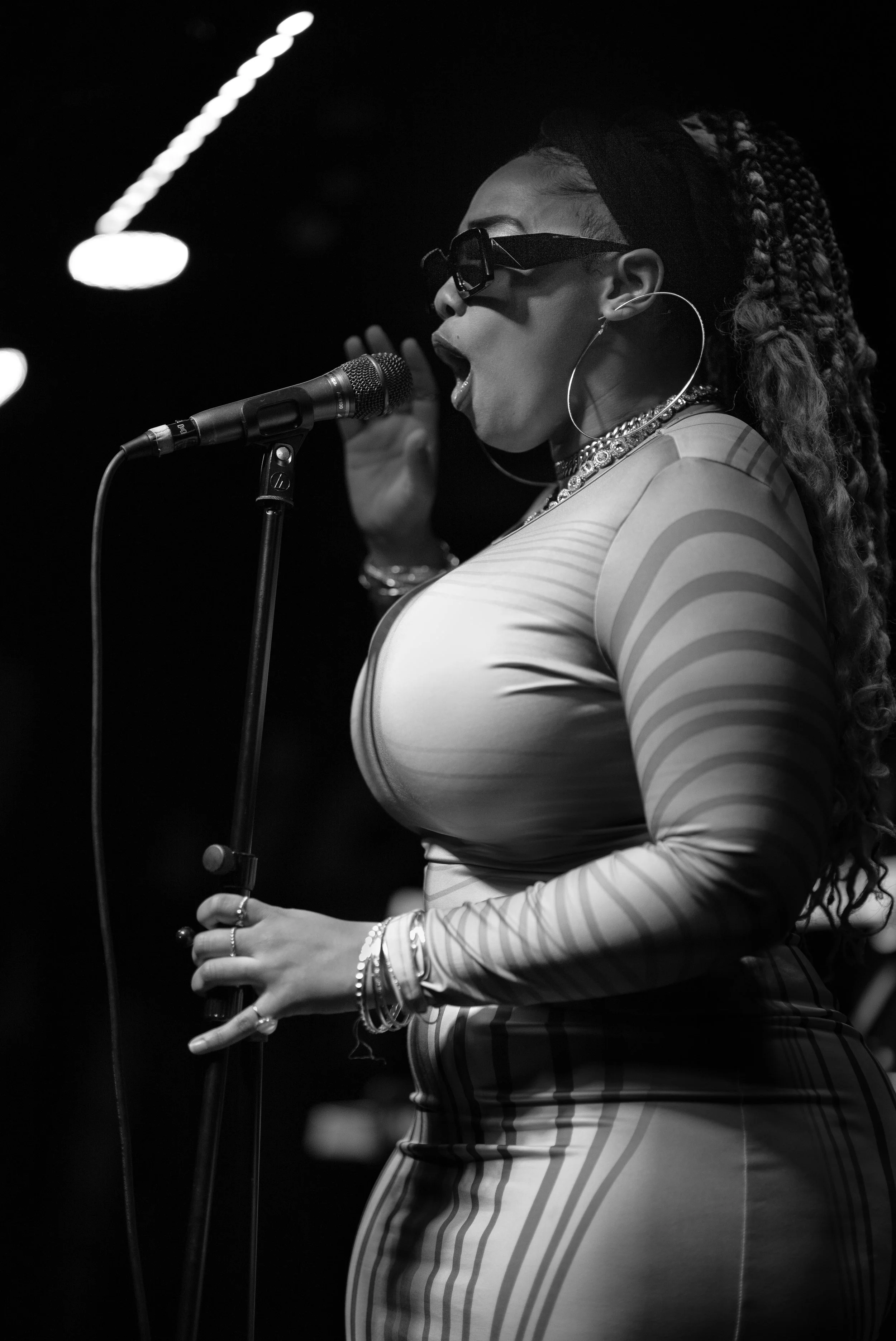 A Black woman singing into a microphone on stage, wearing sunglasses, large hoop earrings, and a fitted, striped top.