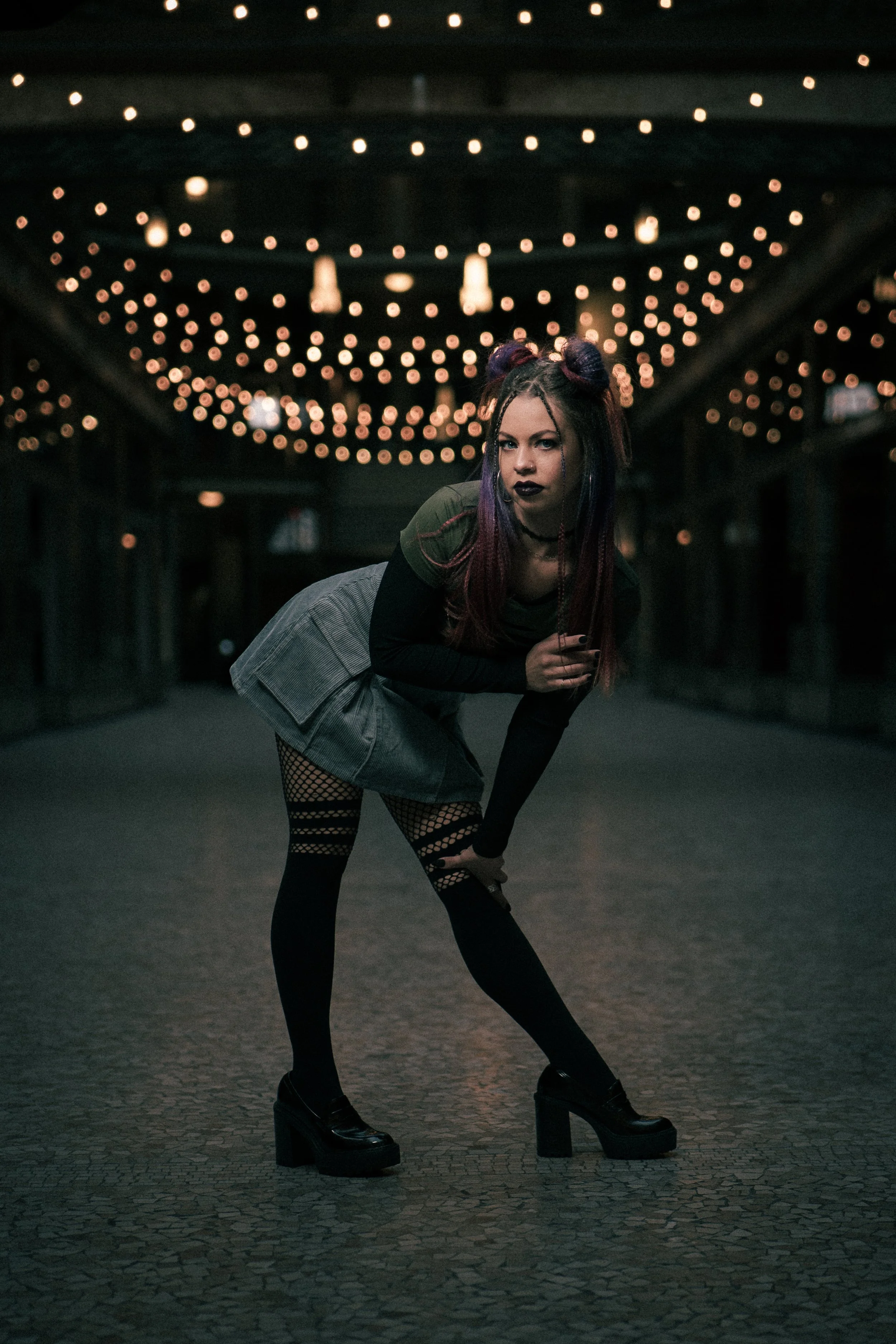 A young woman with purple hair, dressed in gothic style, standing in a dark indoor space with string lights overhead, wearing black platform shoes, a grey skirt, fishnet stockings, and dark makeup.